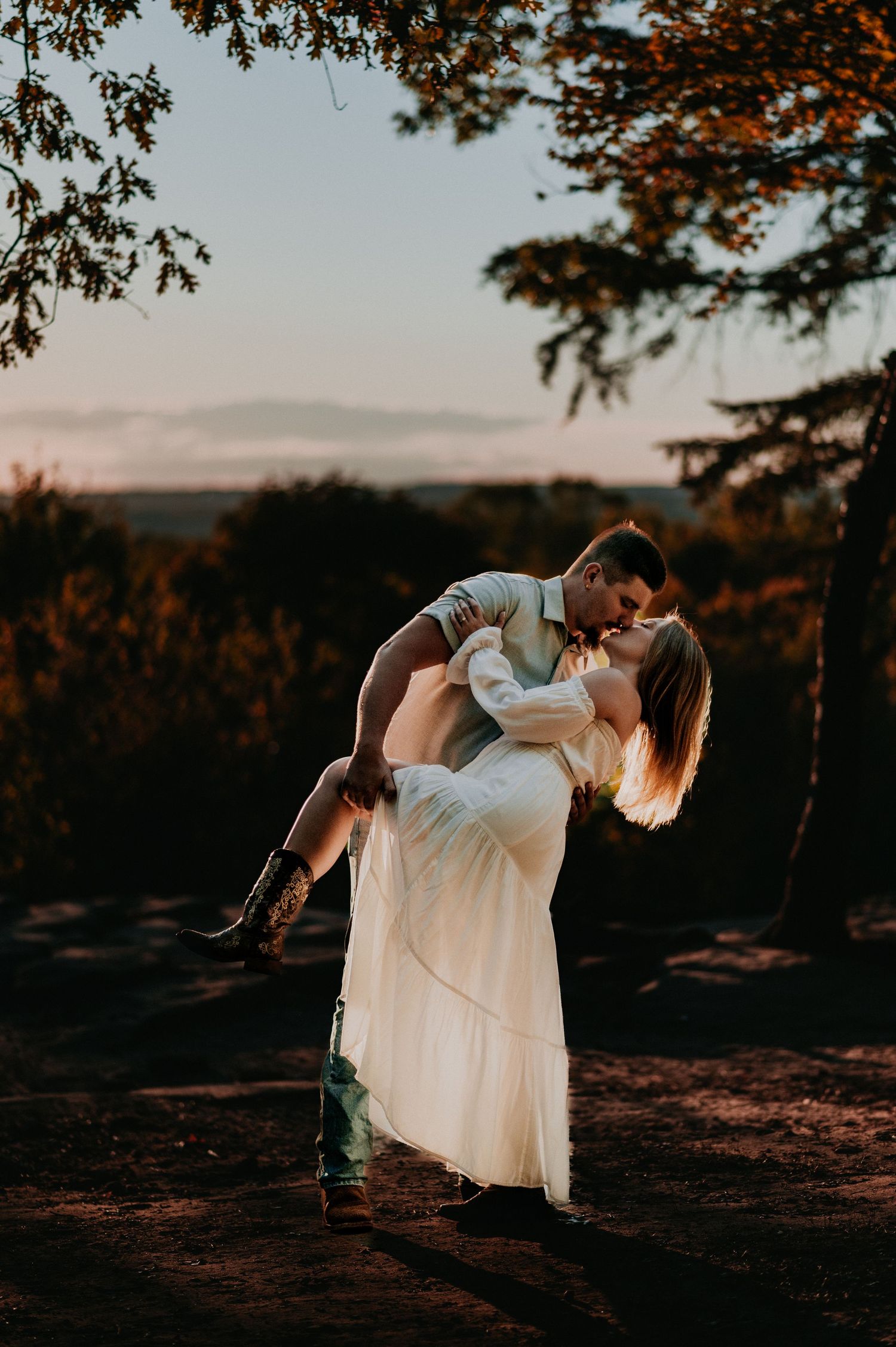 A romantic couple embraces in a white dress and casual wear during sunset in a natural outdoor setting.