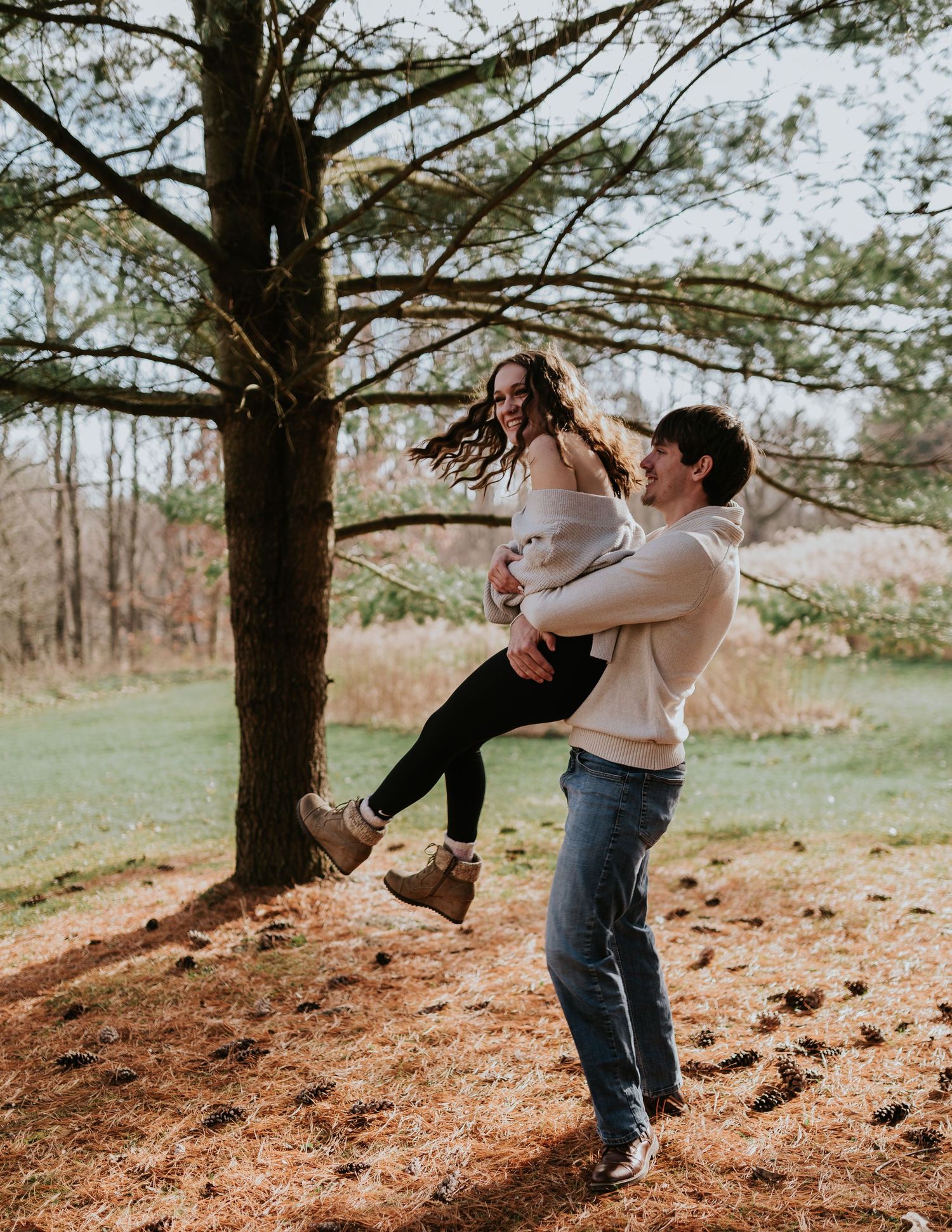 A joyful couple playing and spinning under a pine tree during their engagement session.