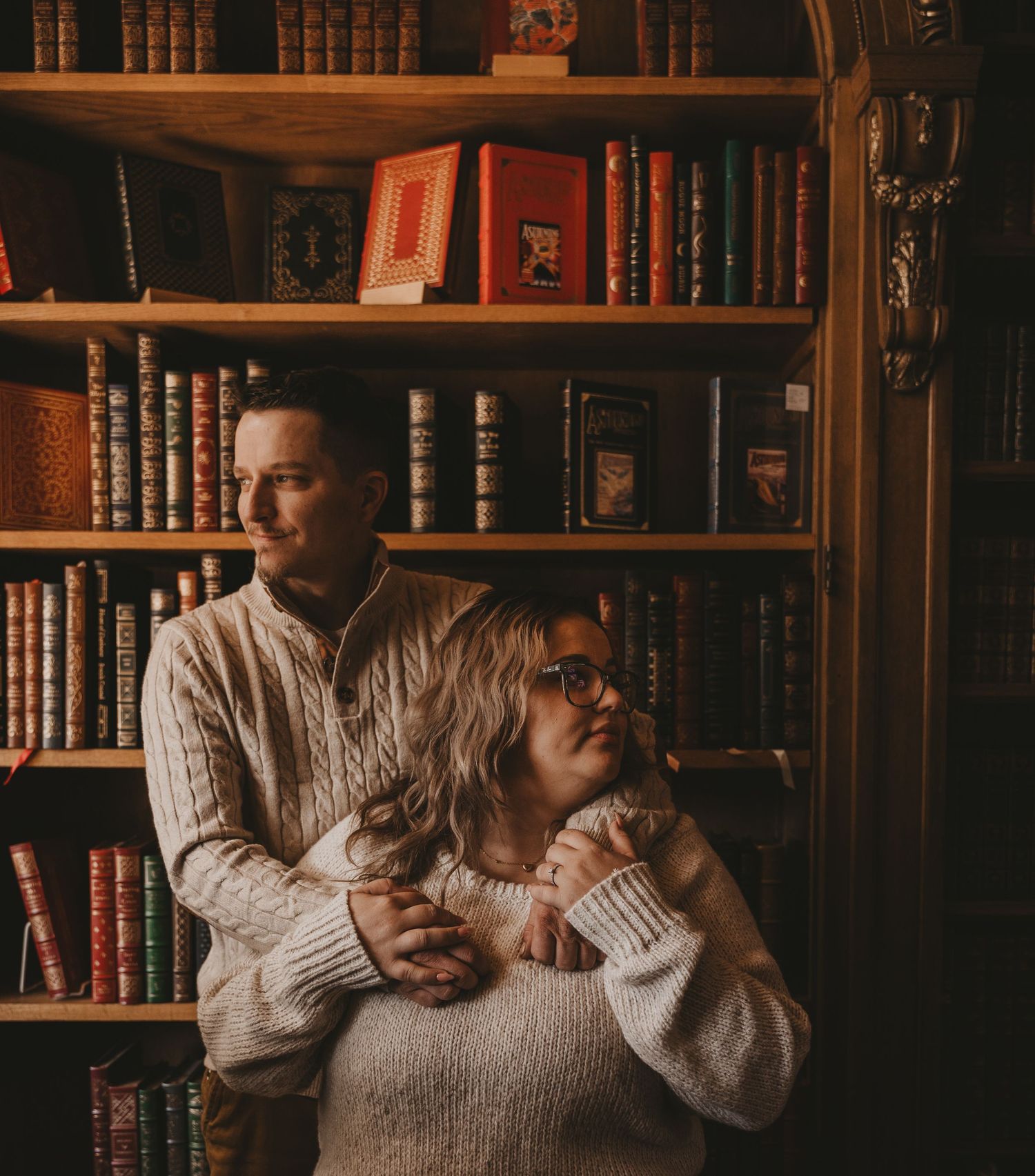 A couple embraces in front of vintage bookshelves in a warmly lit library setting wearing cream sweaters.