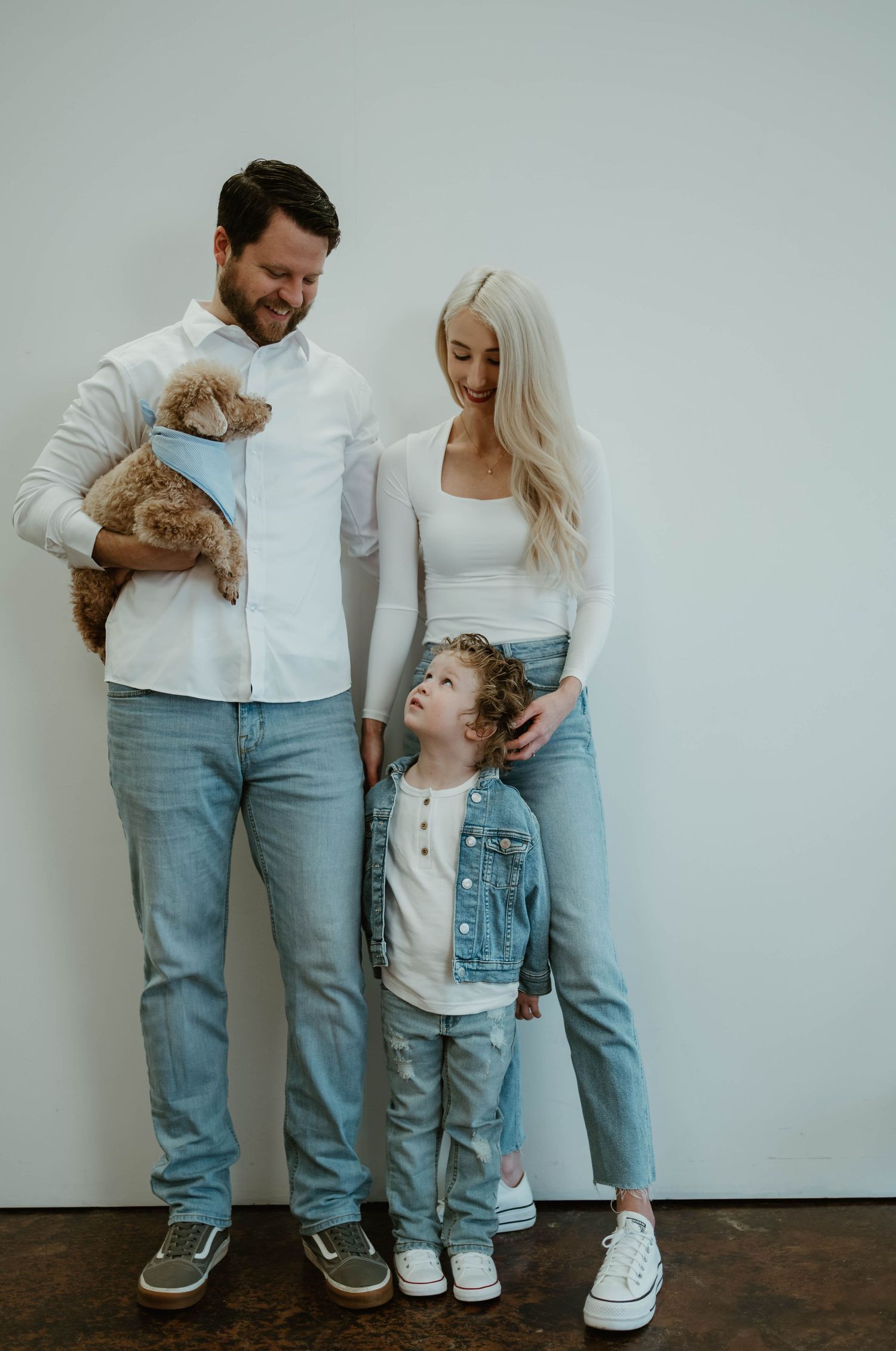 A family poses against a white wall wearing matching denim and white outfits while holding a small ginger cat.