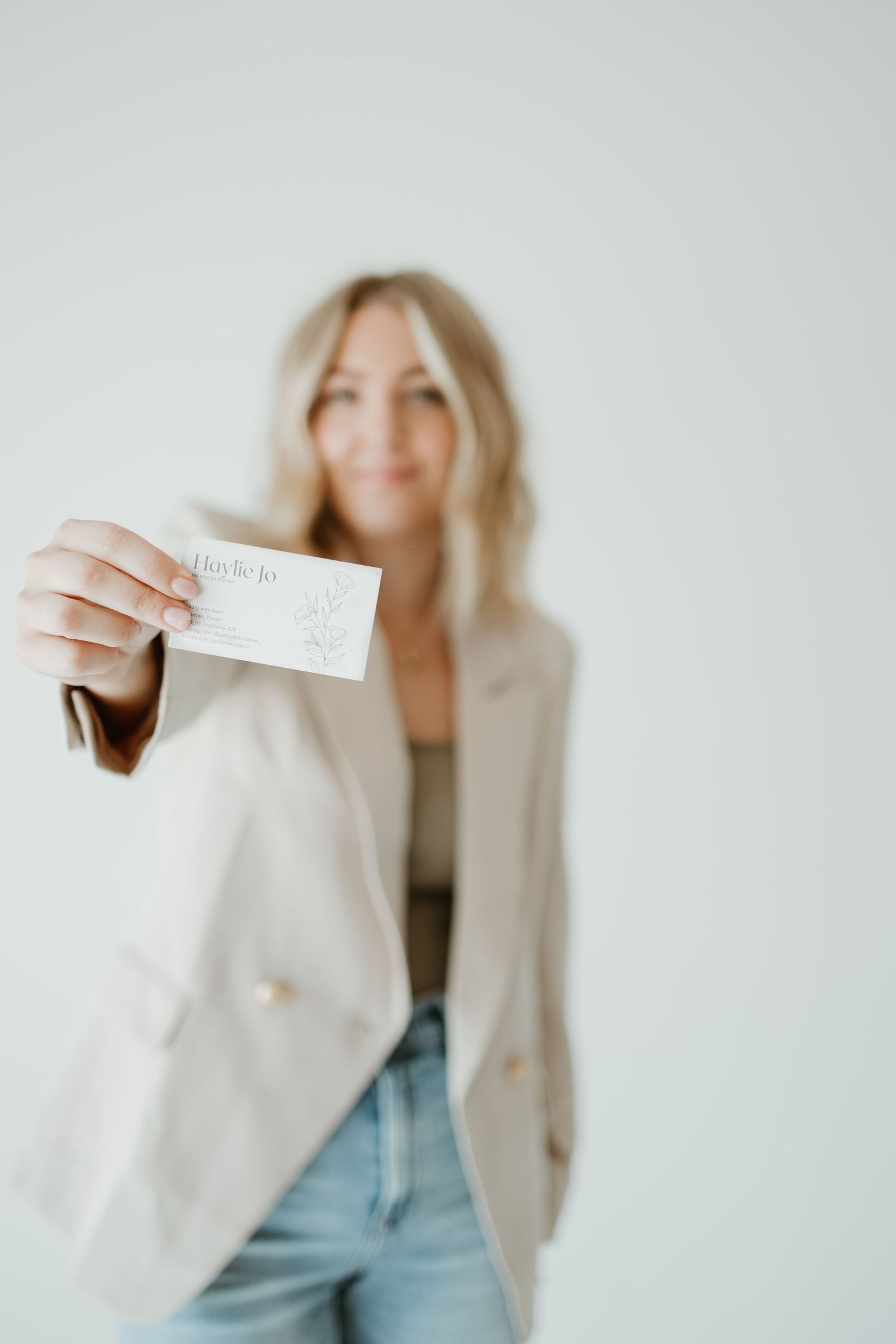 Person in beige blazer and jeans showing business card against light background.