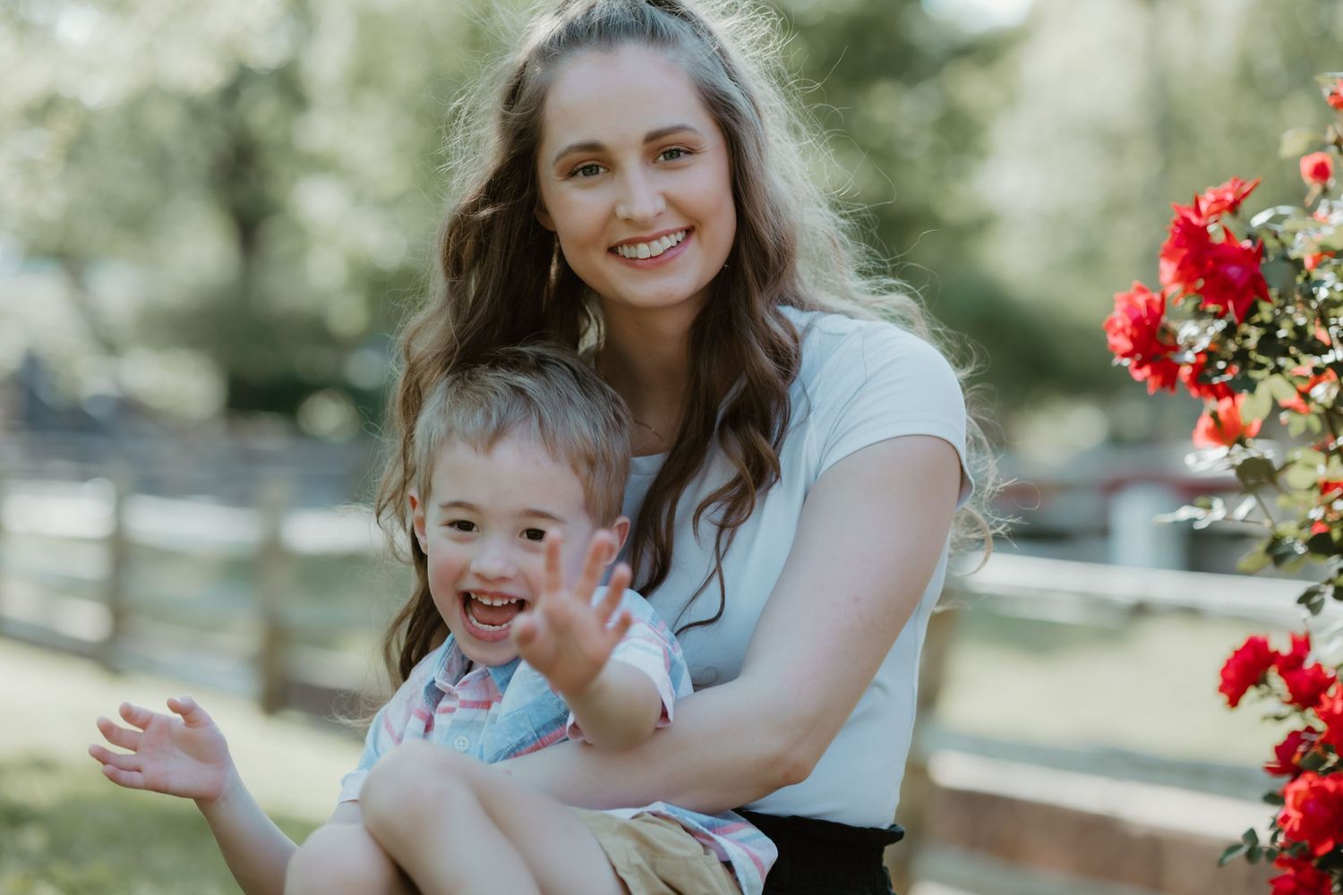 A parent and child sitting outdoors in front of red flowers and wooden fencing on a sunny day.