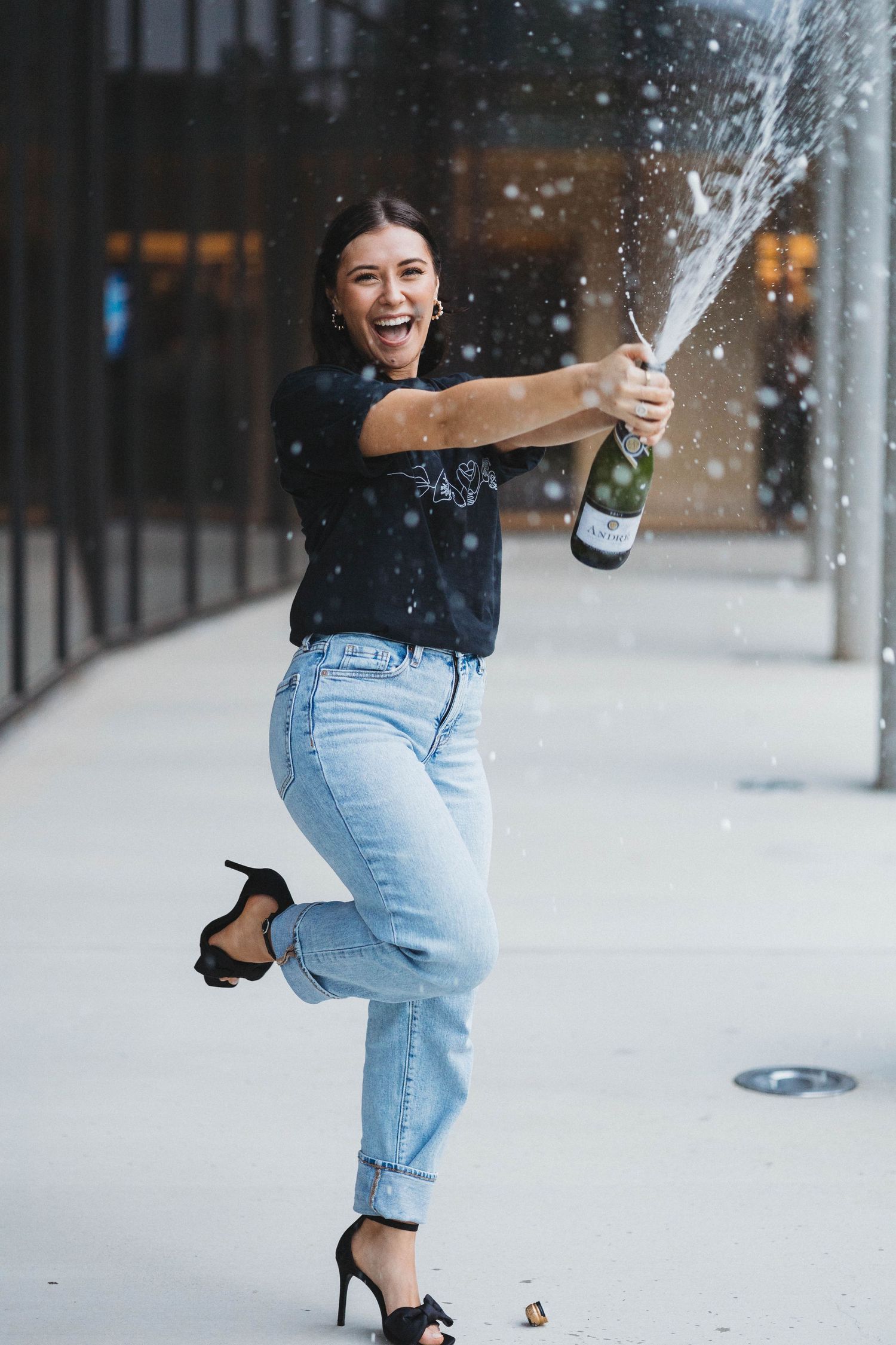Someone celebrating with champagne spray while wearing black top, jeans and heels on city sidewalk.
