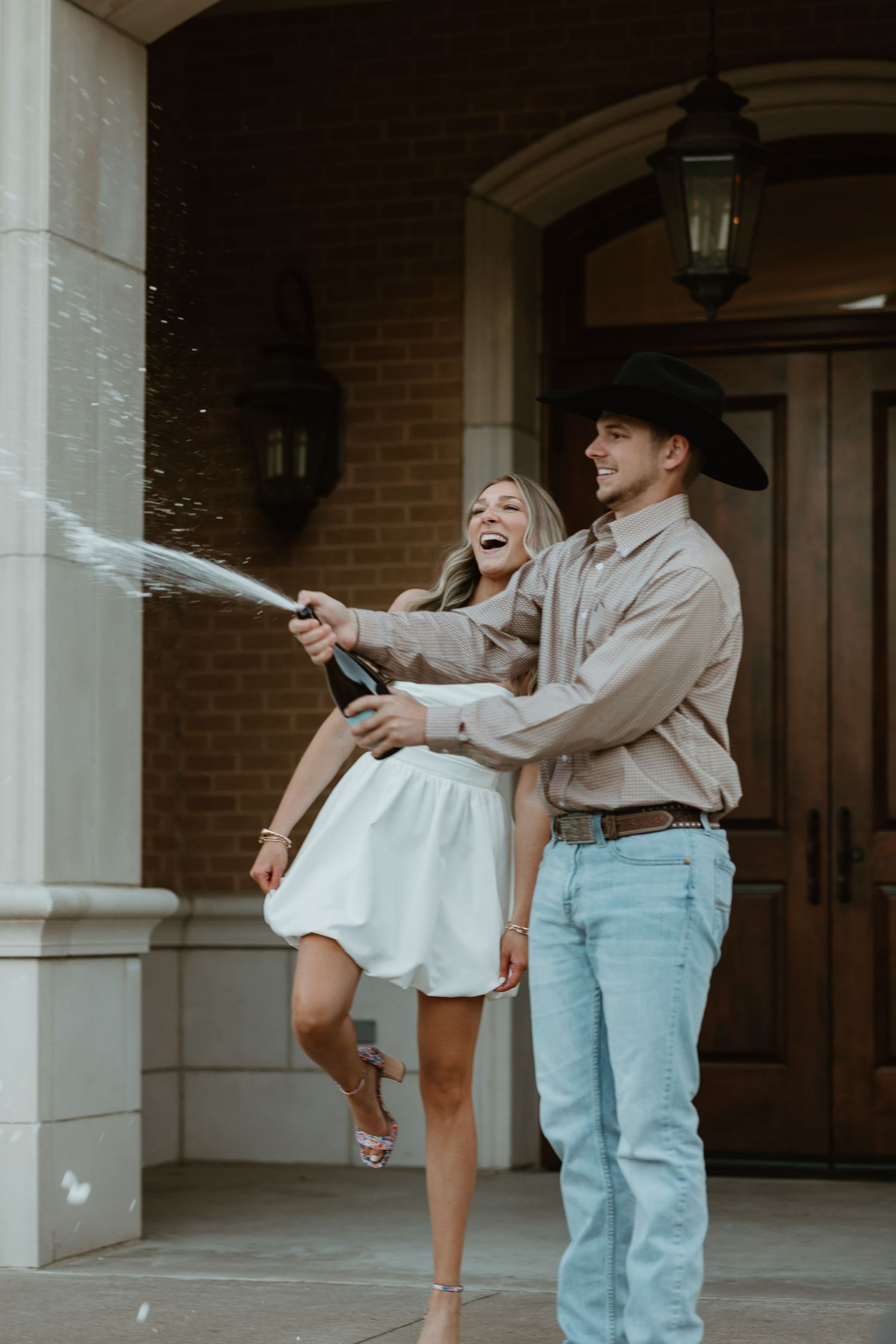 A couple playfully sprays champagne at each other during a celebratory photo shoot at a brick building entrance.