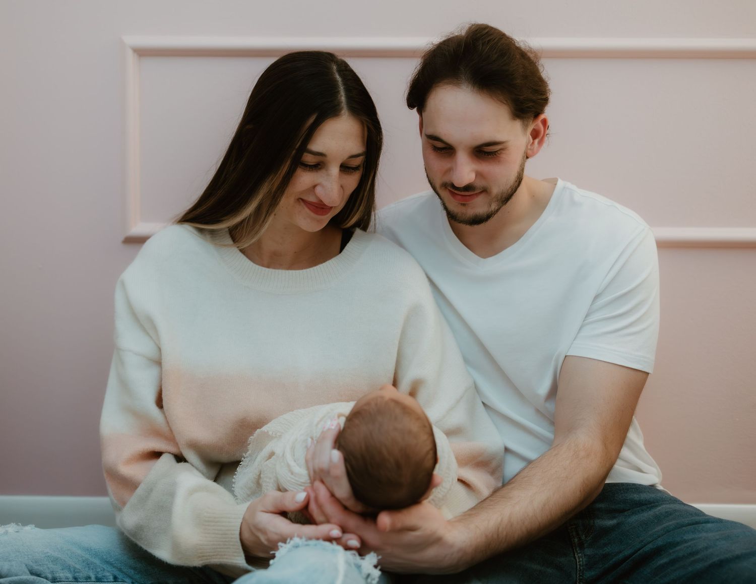 A couple in white tops sits together tenderly looking down at their newborn baby against a light pink wall.