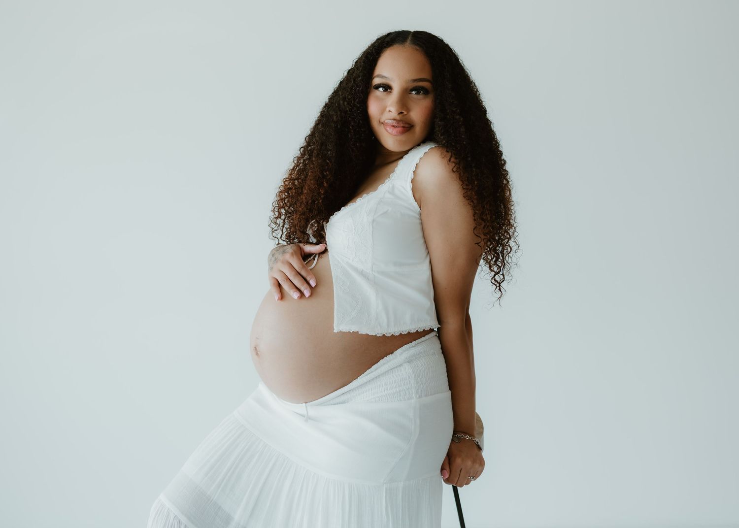 Woman in flowing white bridal dress and long curly hair poses against light backdrop in minimalist studio setting.