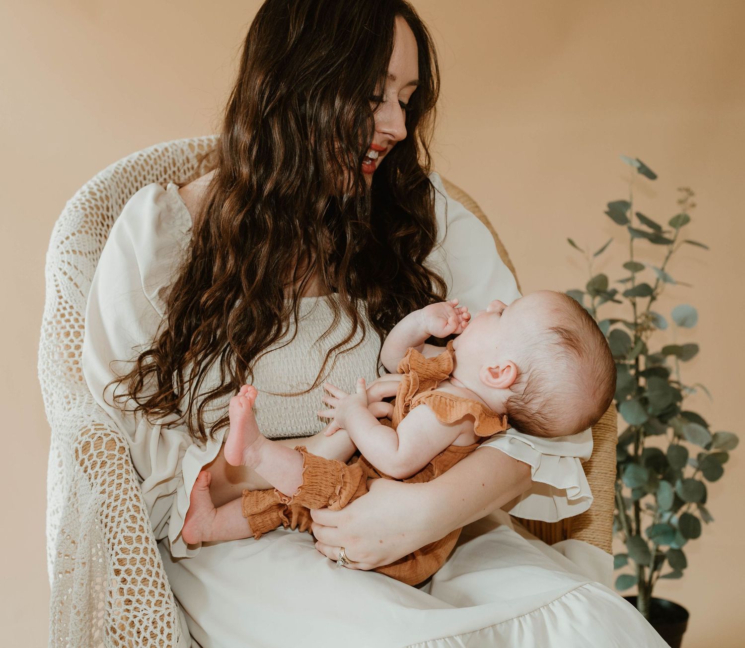 A person in a white dress holds a sleeping infant in a serene moment with a eucalyptus plant backdrop.