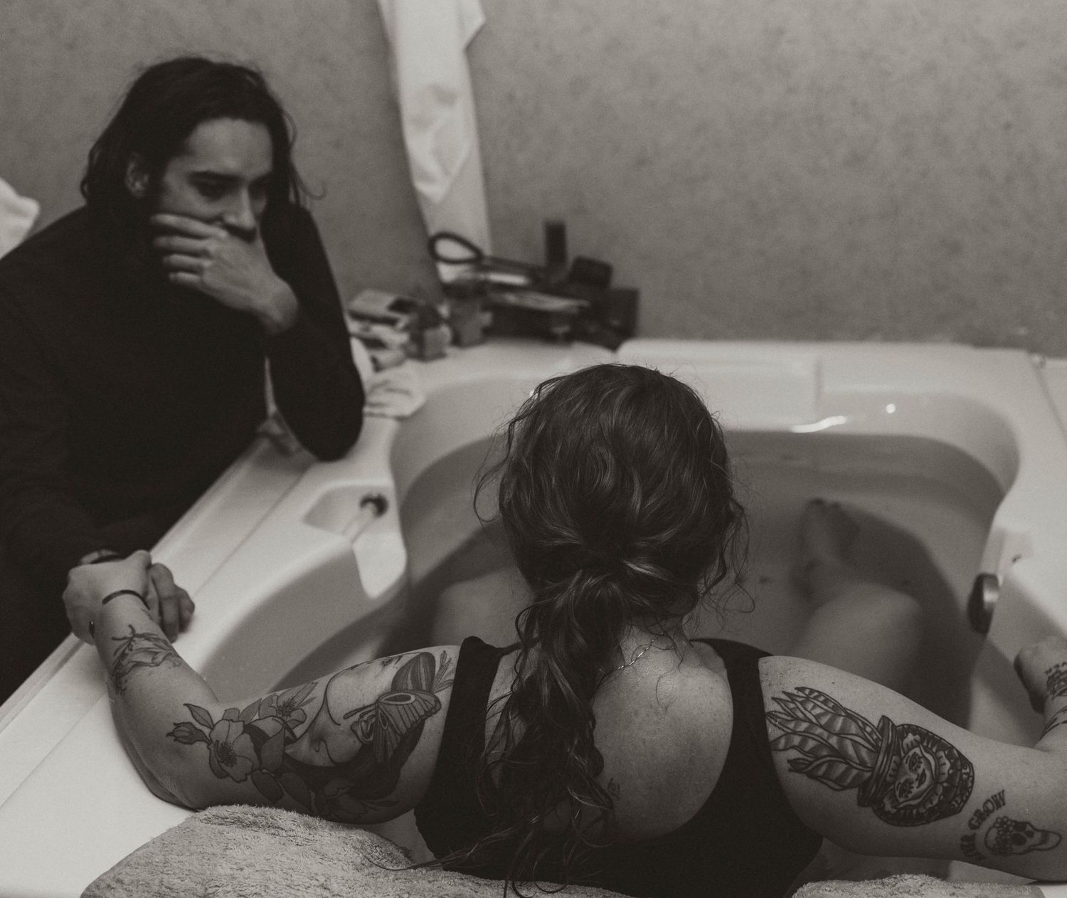 Person giving birth in white bathtub with long black hair wearing white clothing and head resting on edge.
