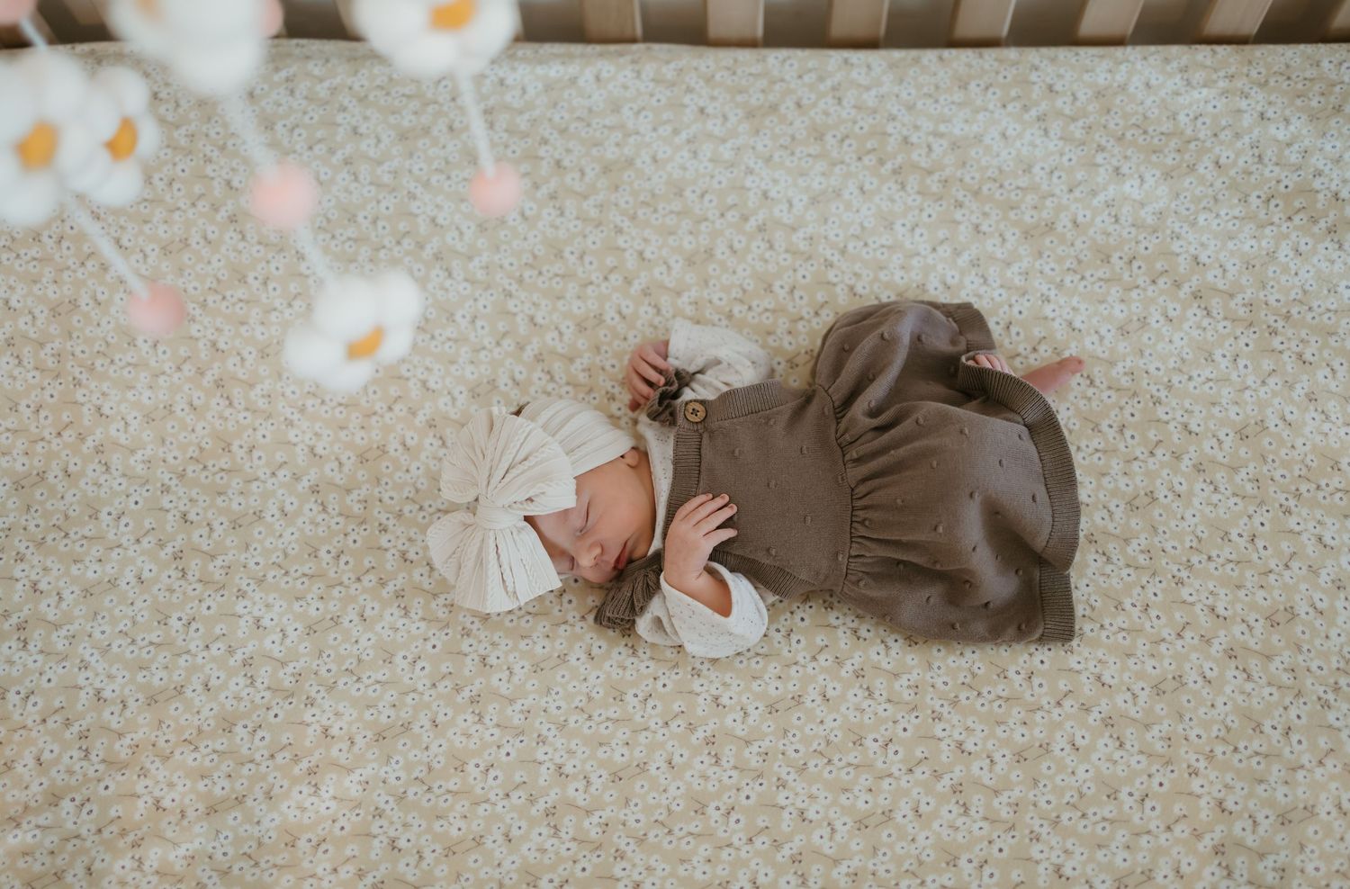 Baby wearing brown dress lies on carpeted floor surrounded by soft pastel flower petals.