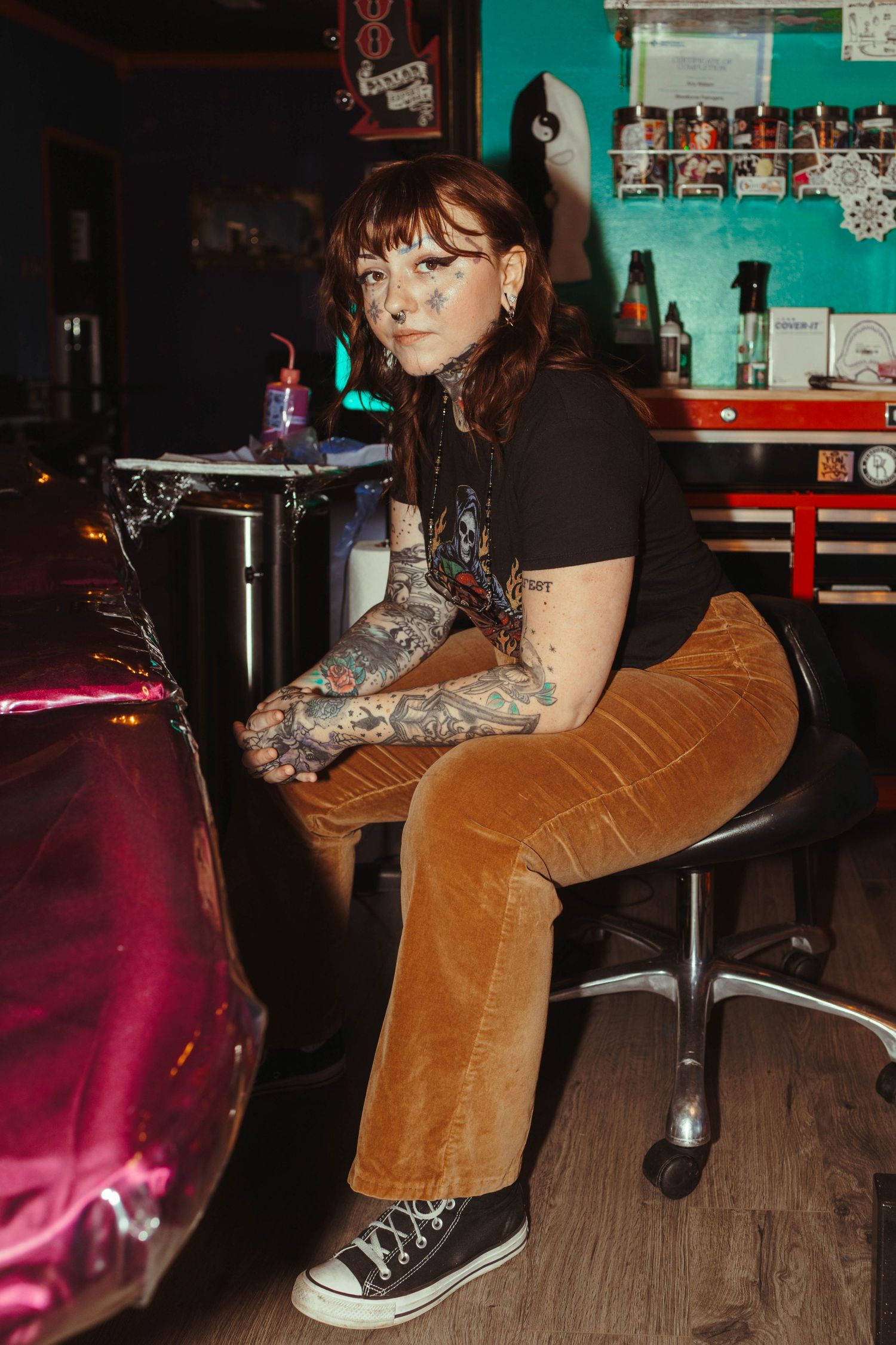 Someone in brown corduroy pants and black shoes sits on a wooden barstool at a dimly lit venue.