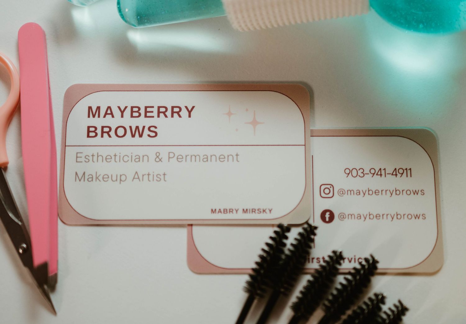 A business card for Mayberry Brows makeup artist alongside pink tools and black mascara wands on a teal background.