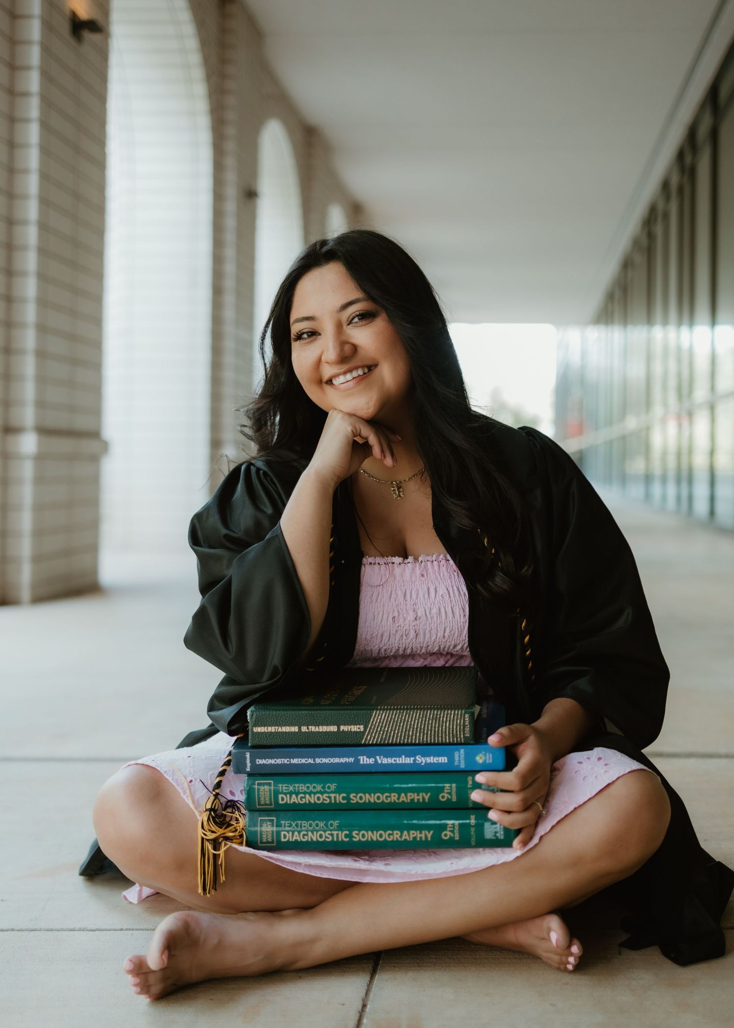 Student sits cross-legged in a hallway with a stack of diagnostic radiology textbooks while wearing pink top and black jacket.