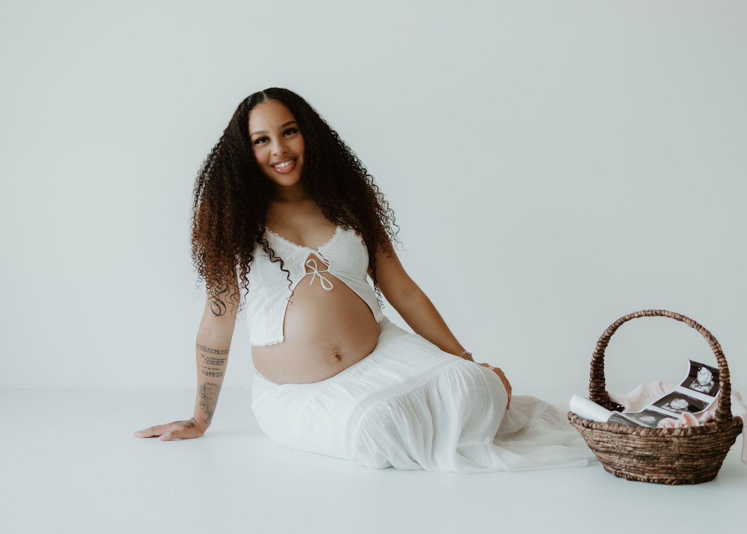 Woman wearing white dress poses gracefully against light gray backdrop with woven basket.