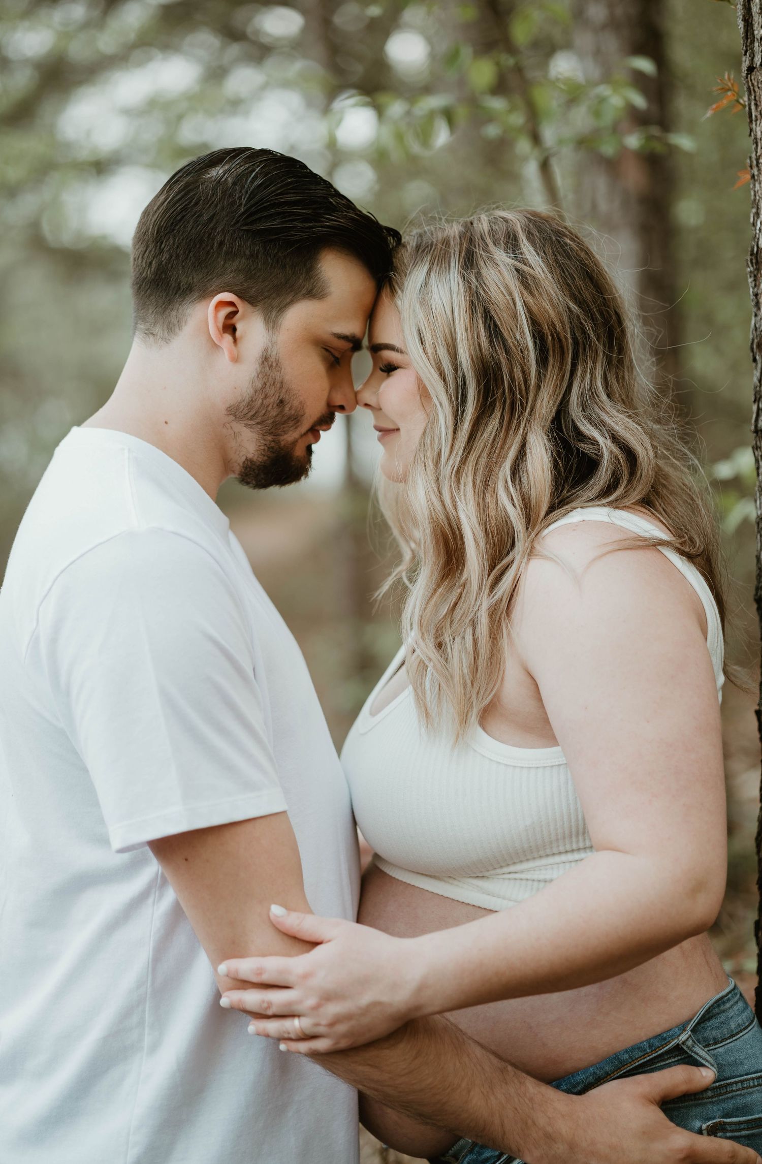 A romantic couple in white shares an intimate forehead touch while embracing in a softly lit forest setting.