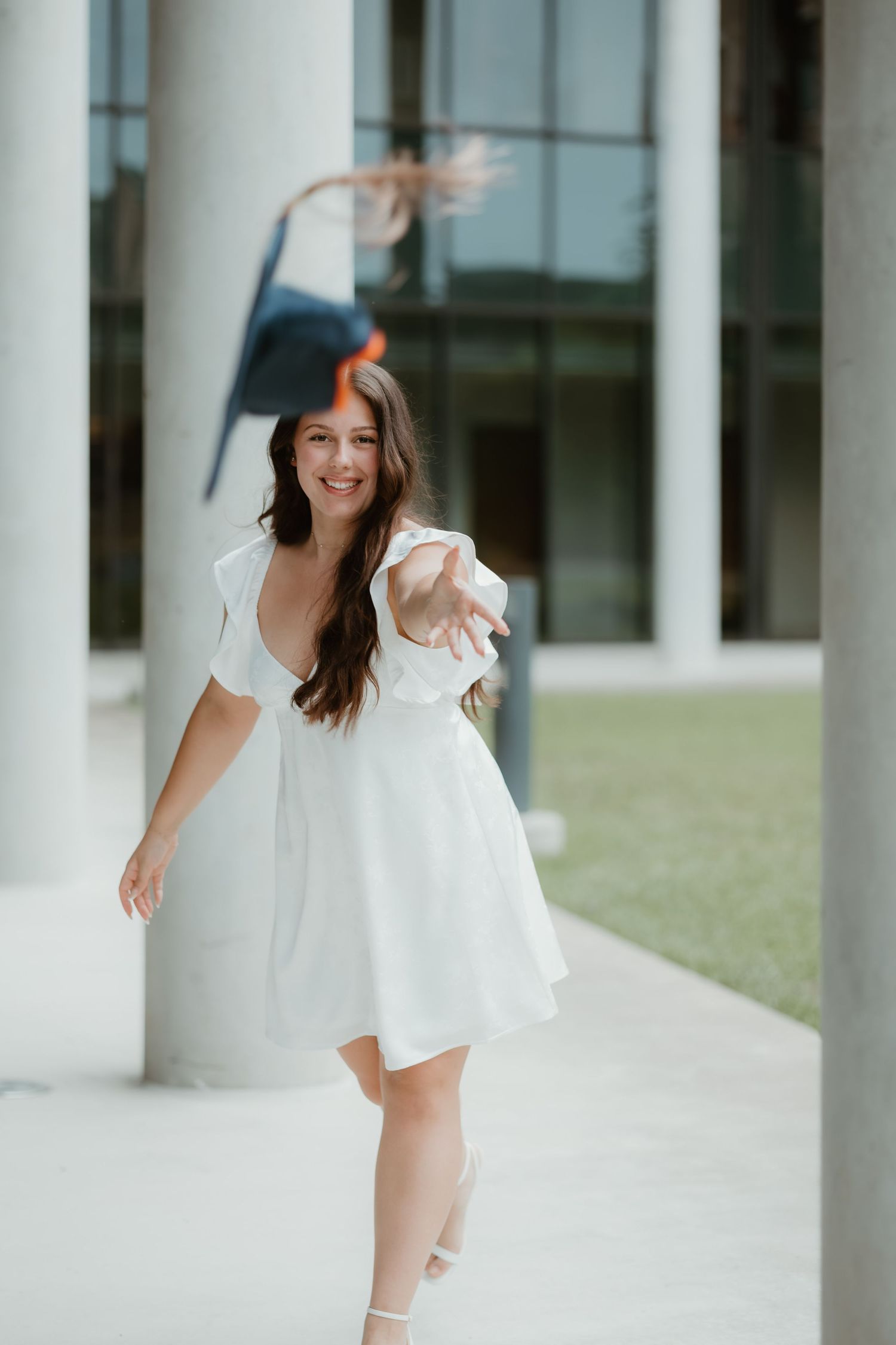 Graduate in white dress celebrates outdoors on campus with graduation cap during sunny graduation photoshoot.
