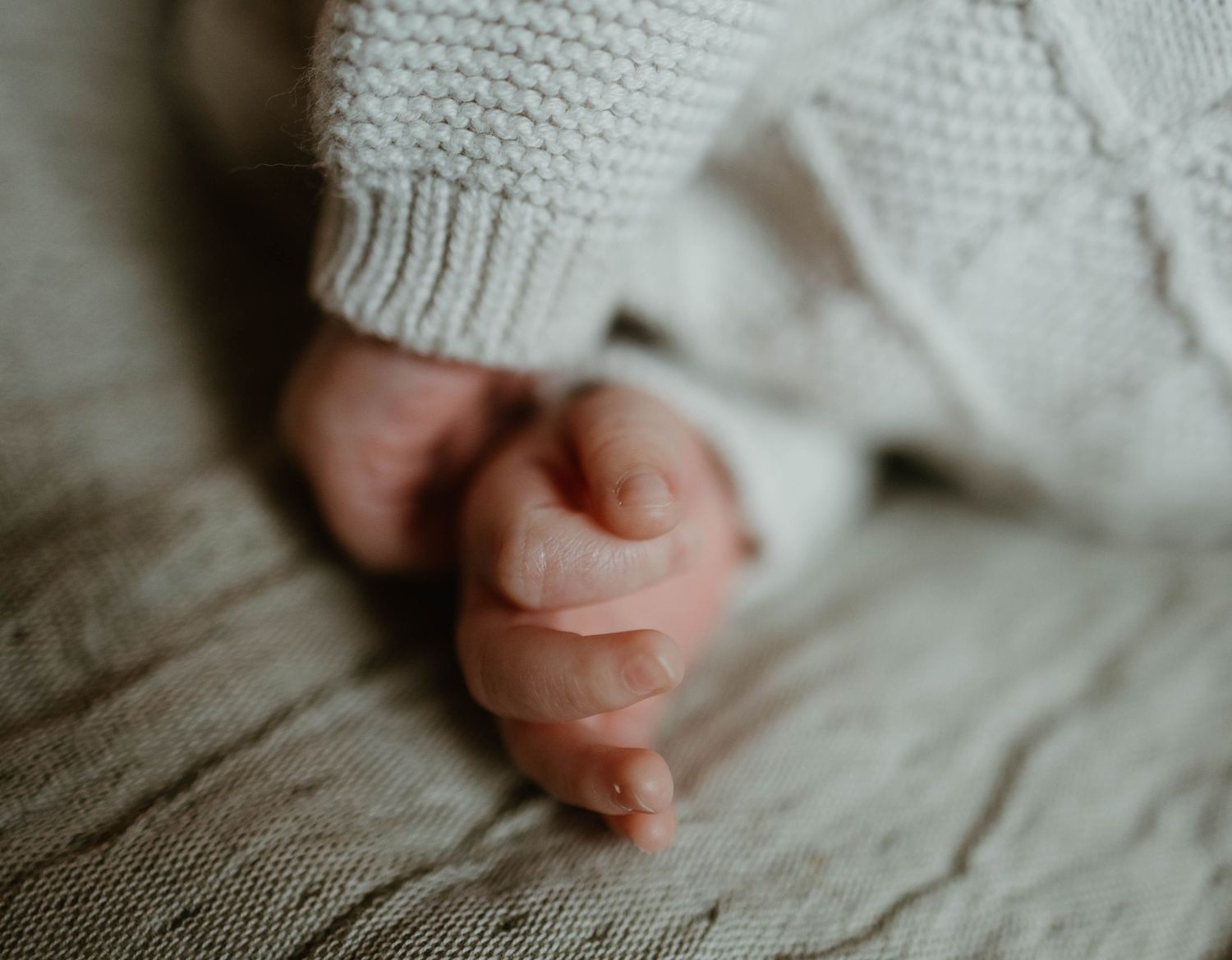 Tiny baby foot in cozy white knit sweater rests on soft gray blanket in natural lighting.