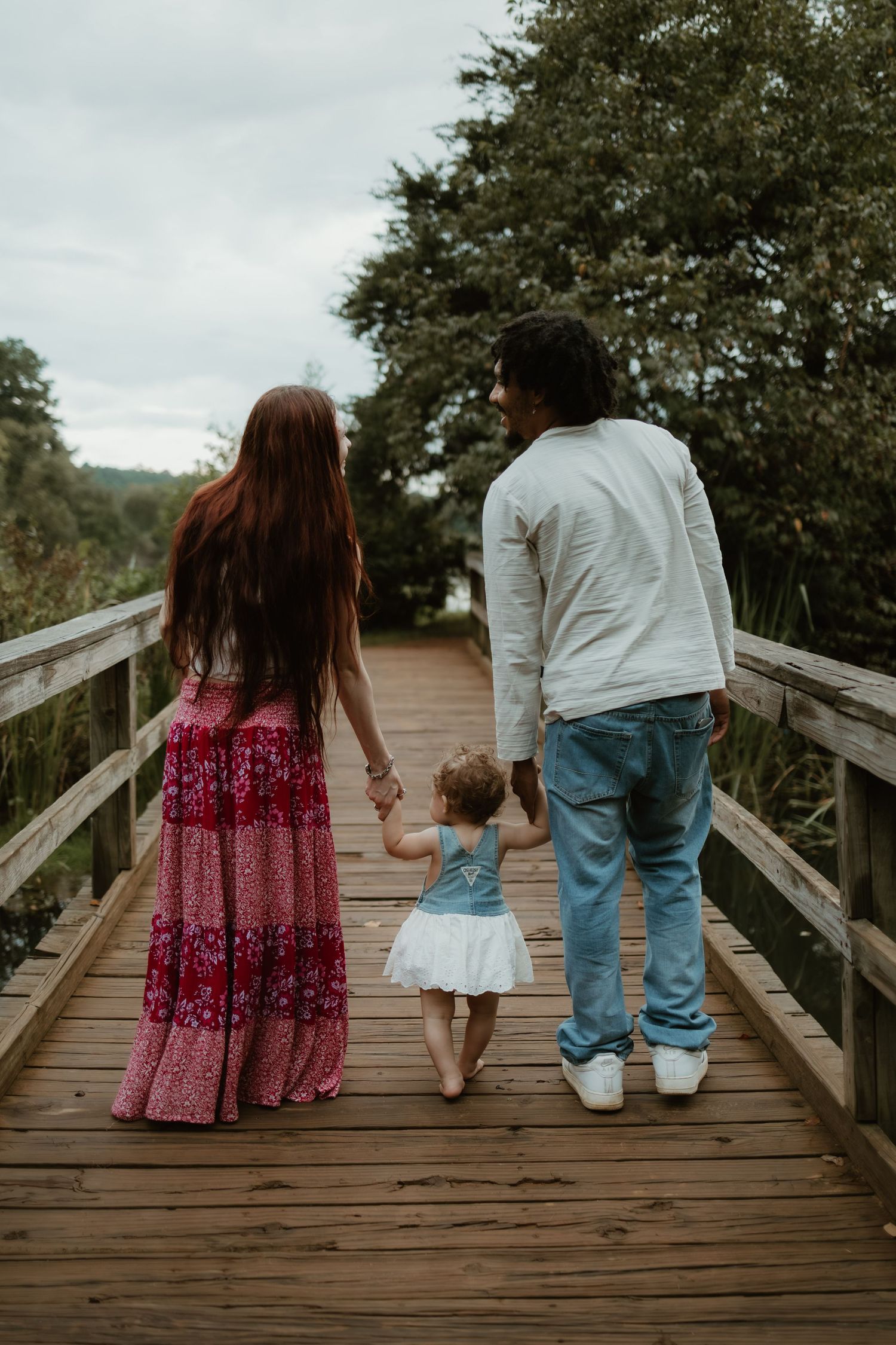 Two individuals in casual attire walk together on a wooden boardwalk surrounded by green trees on a cloudy day.