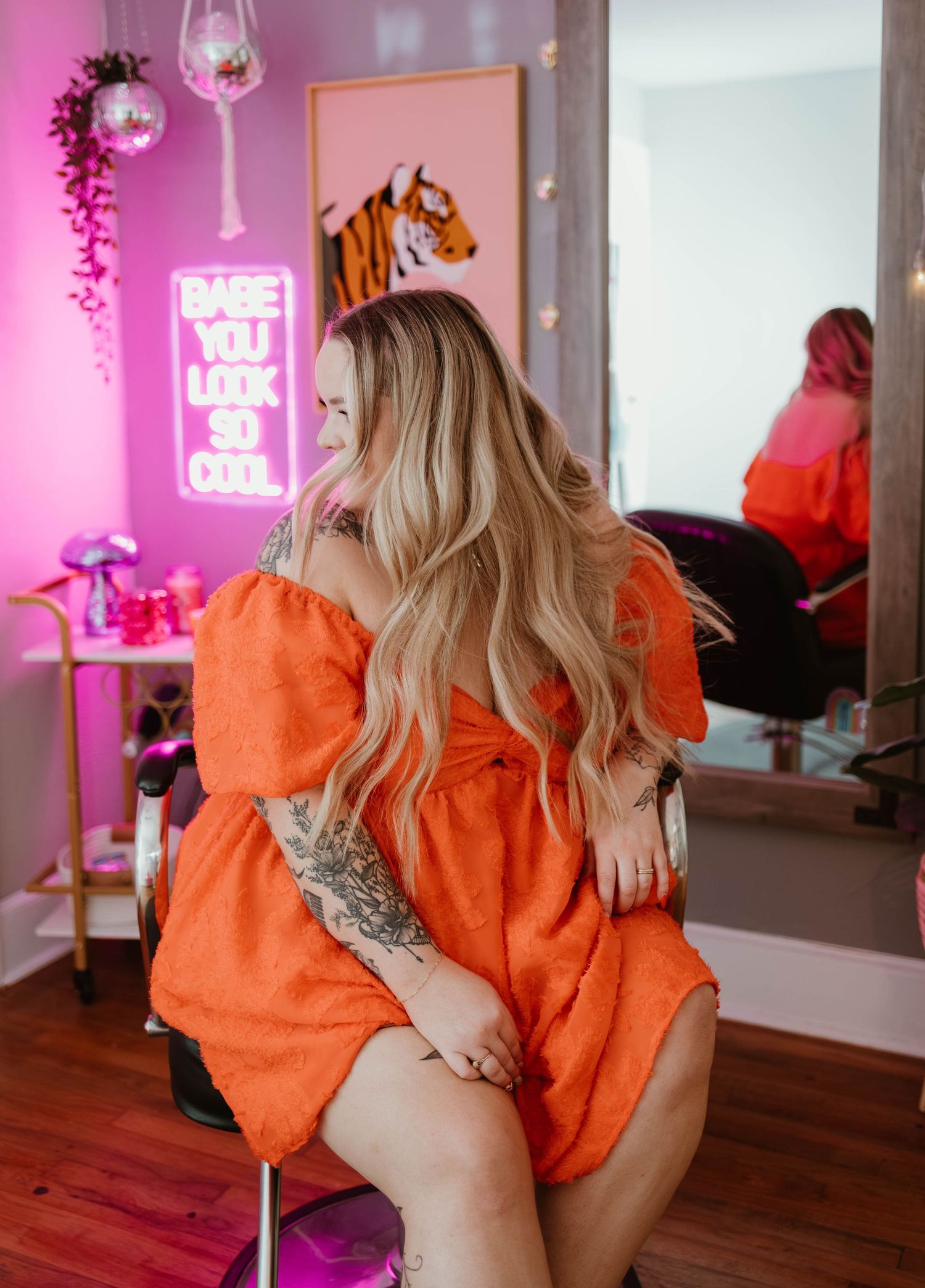 A room with pink neon lighting features a bright orange dress and mirror reflection against warm wood flooring.