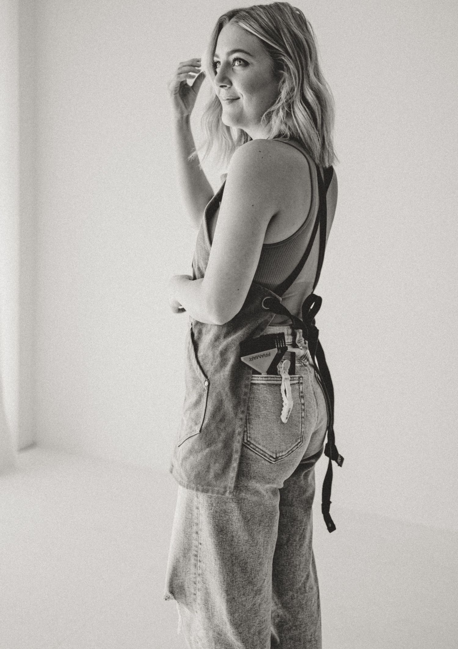 Black and white portrait of someone in denim overalls and tank top posing against white wall.