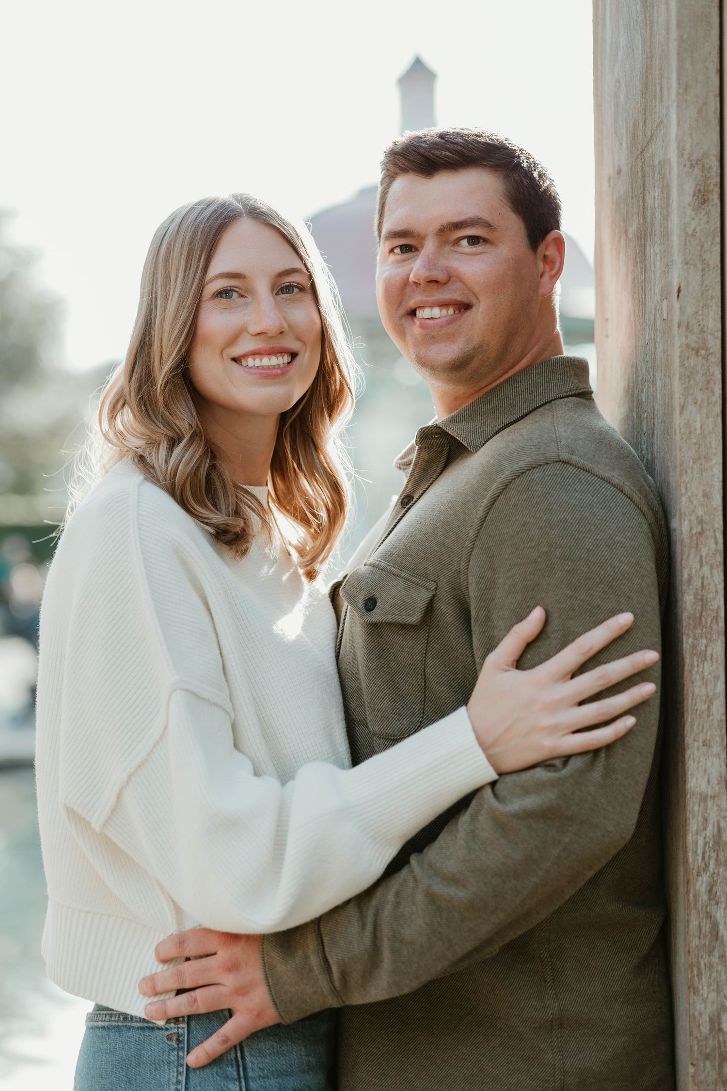 A couple in winter attire embraces outdoors while smiling and showing affection in a romantic engagement photoshoot.