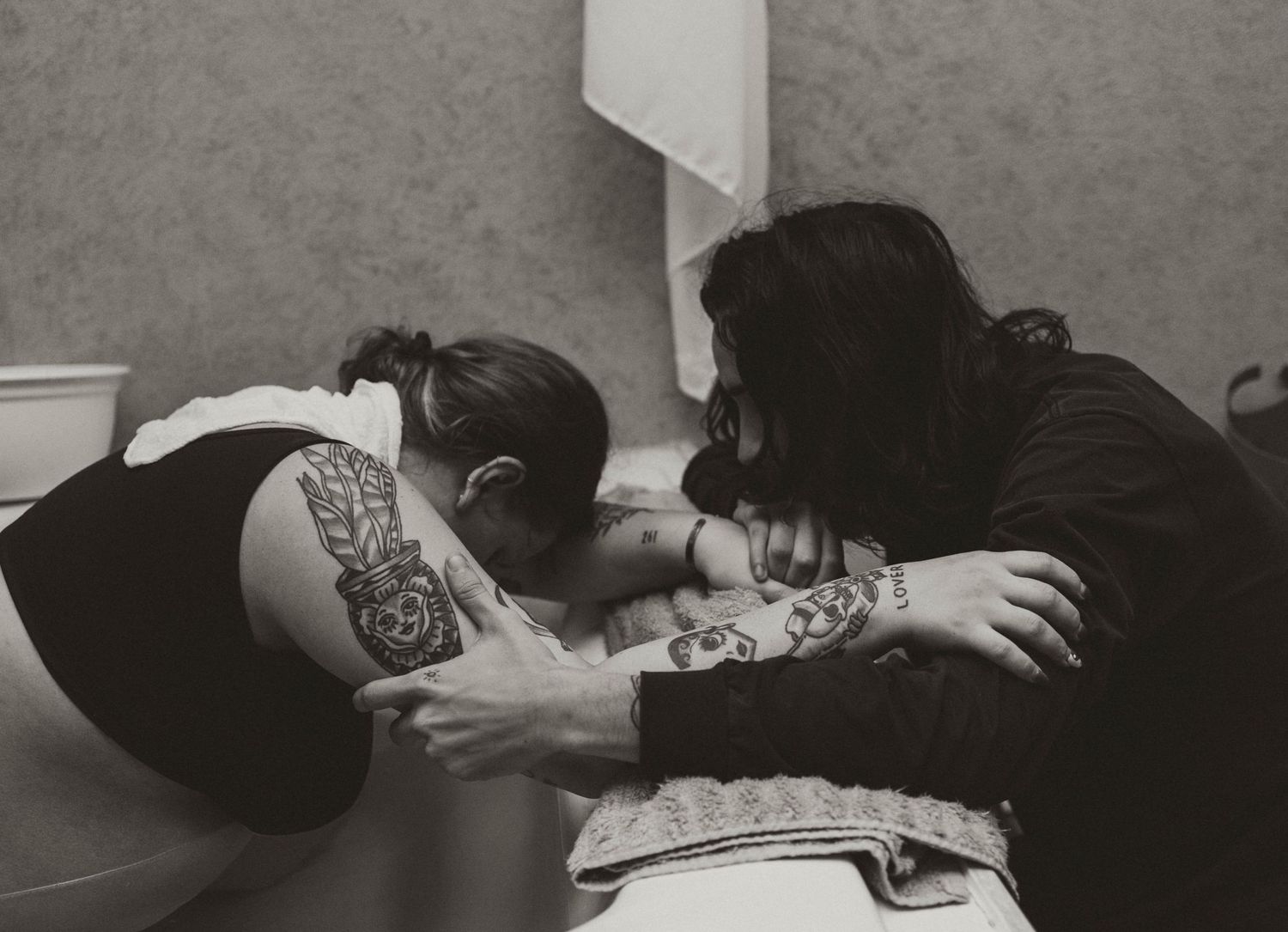 A black and white photograph shows a tattoo artist at work on a client's arm in a traditional studio setting.