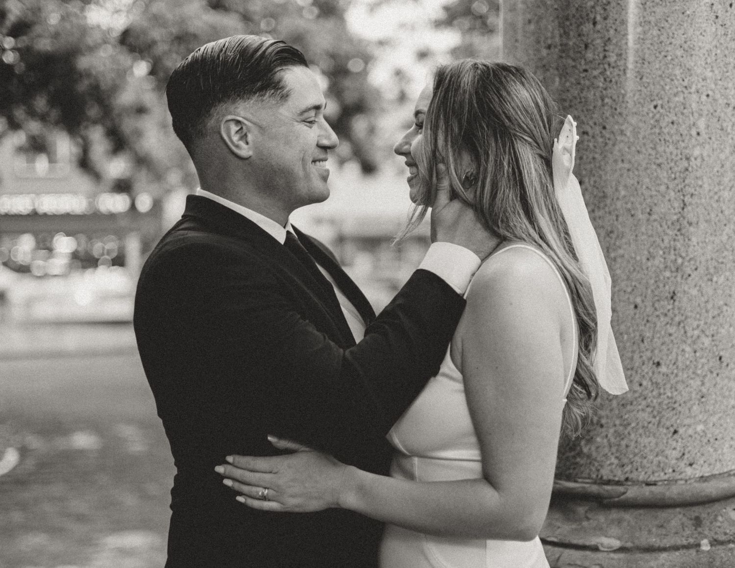 A romantic black and white wedding photograph of a couple embracing by a classic stone column.