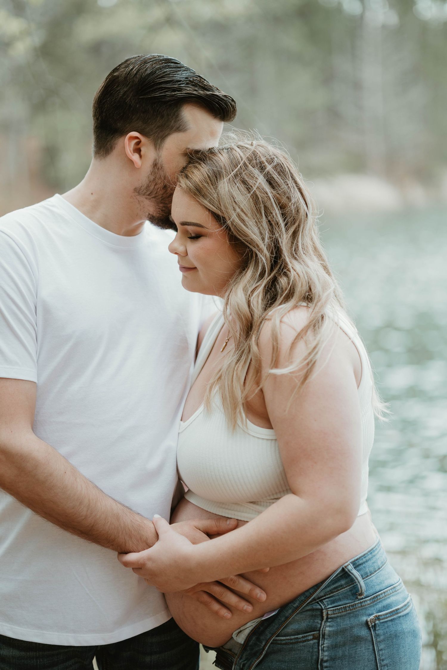 A romantic couple embraces by the water on a soft cloudy day, both wearing casual white tops and sharing an intimate moment.