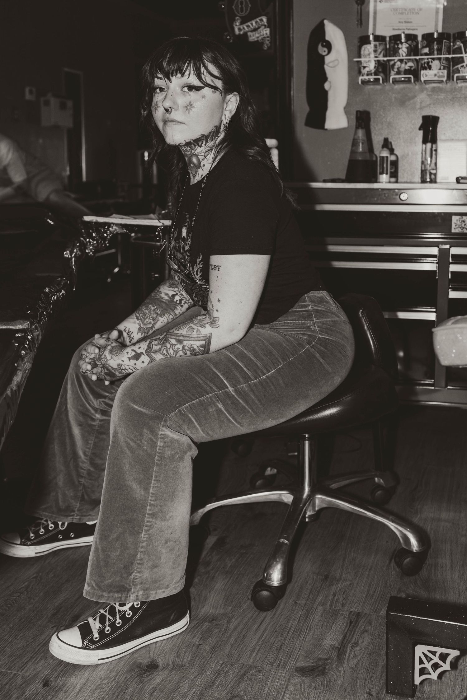Black and white photo of a person with tattoos and glasses sitting on a chair at a bar wearing jeans and Converse sneakers.