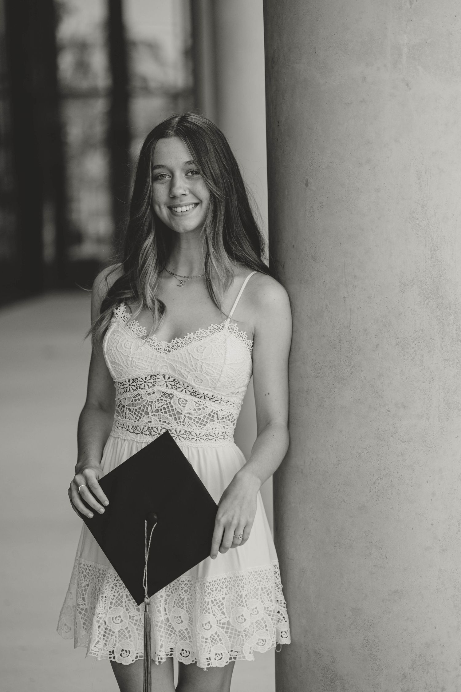 A graduate in a white lace dress holds a diploma while posing against a pillar in black and white photography.