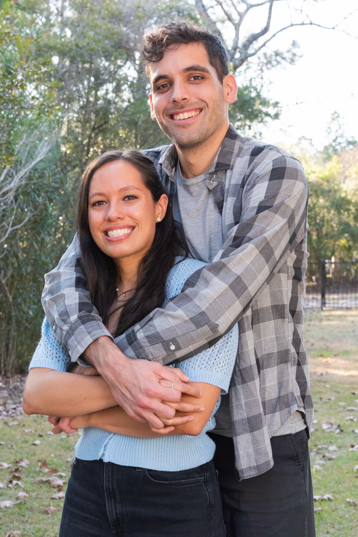 A couple embraces outdoors in a park setting, with the person in a plaid flannel shirt hugging from behind.