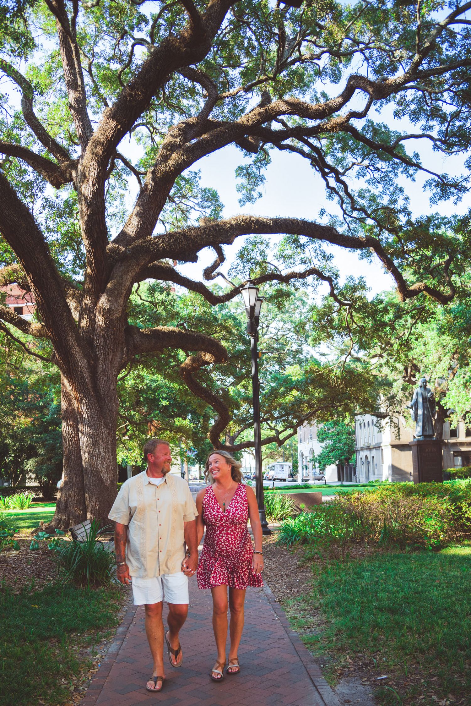 Couple walking hand in hand on a brick path beneath sprawling oak trees in a lush green park.