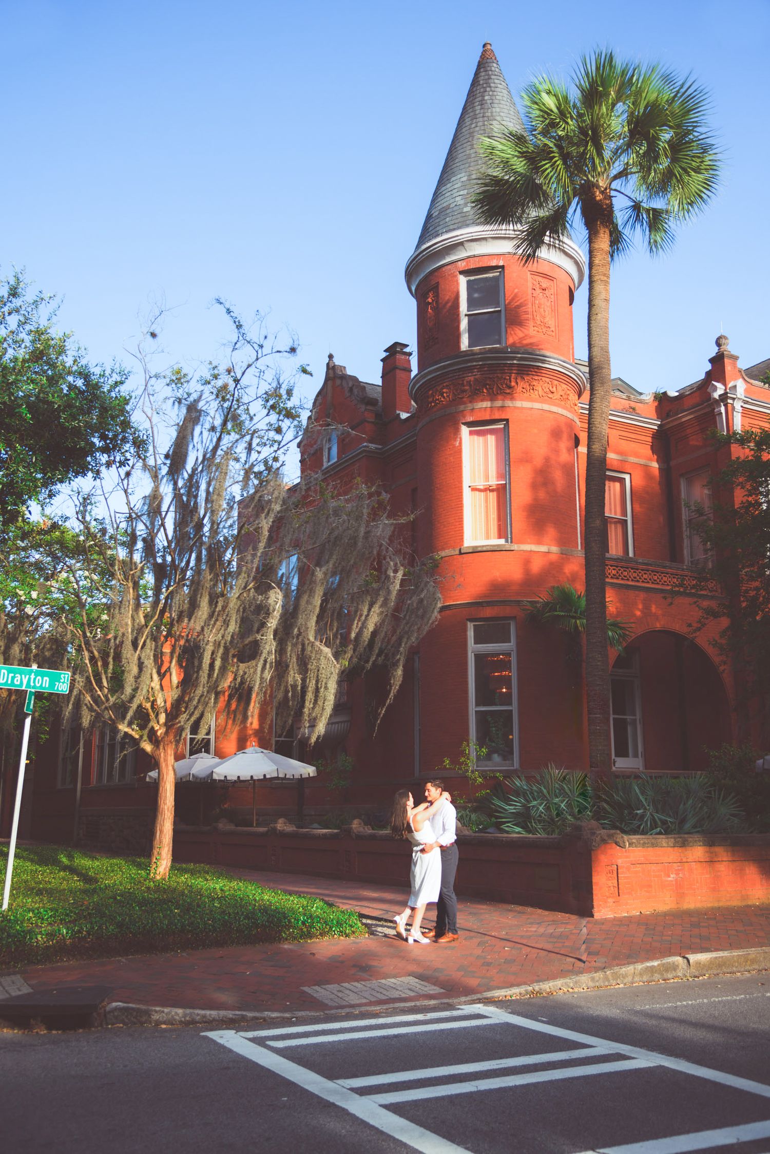 Couple embracing in front of a red brick Victorian building with a turret and palm tree in Savannah, GA.