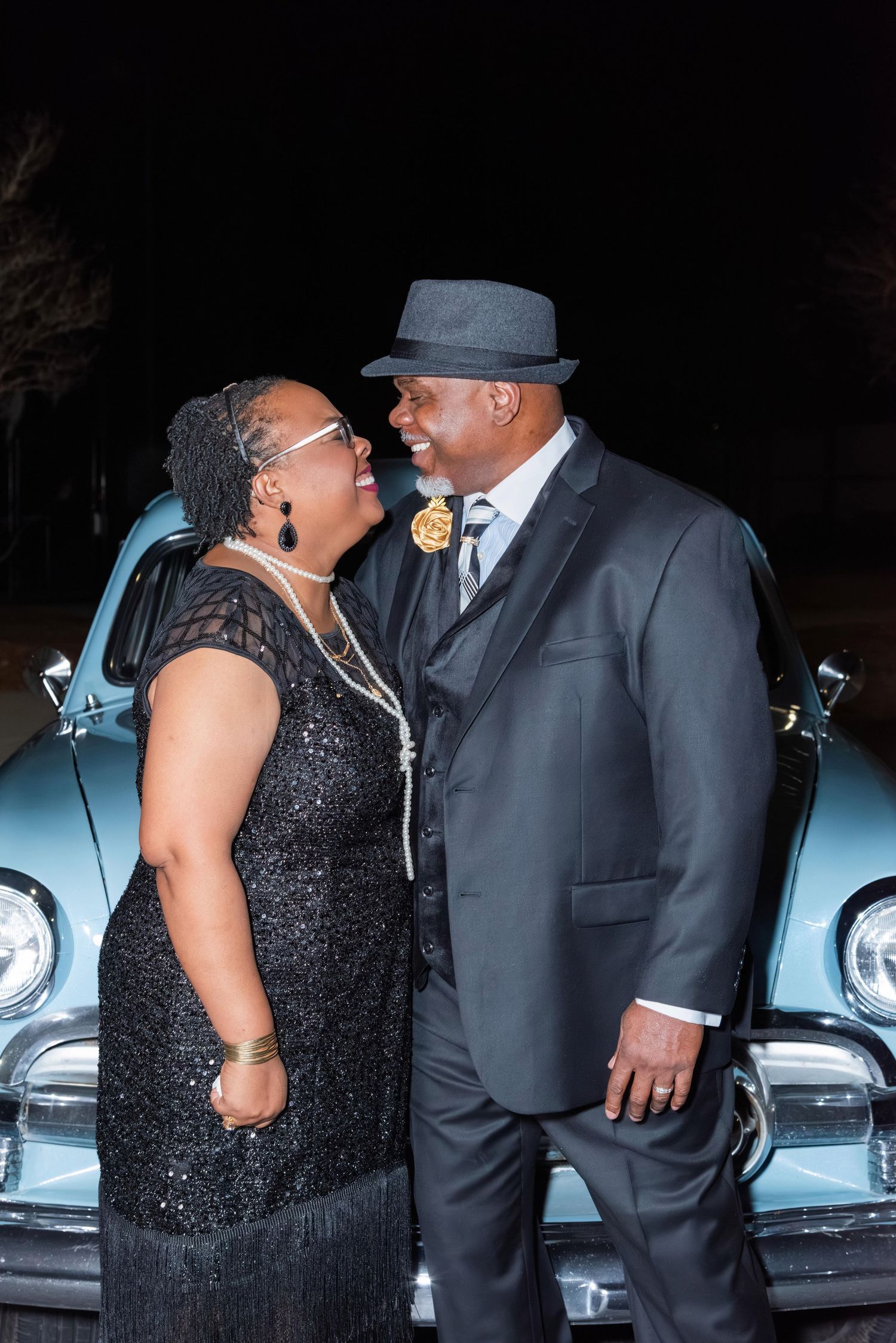 A couple in formal attire sharing a romantic moment in front of a vintage car at night.