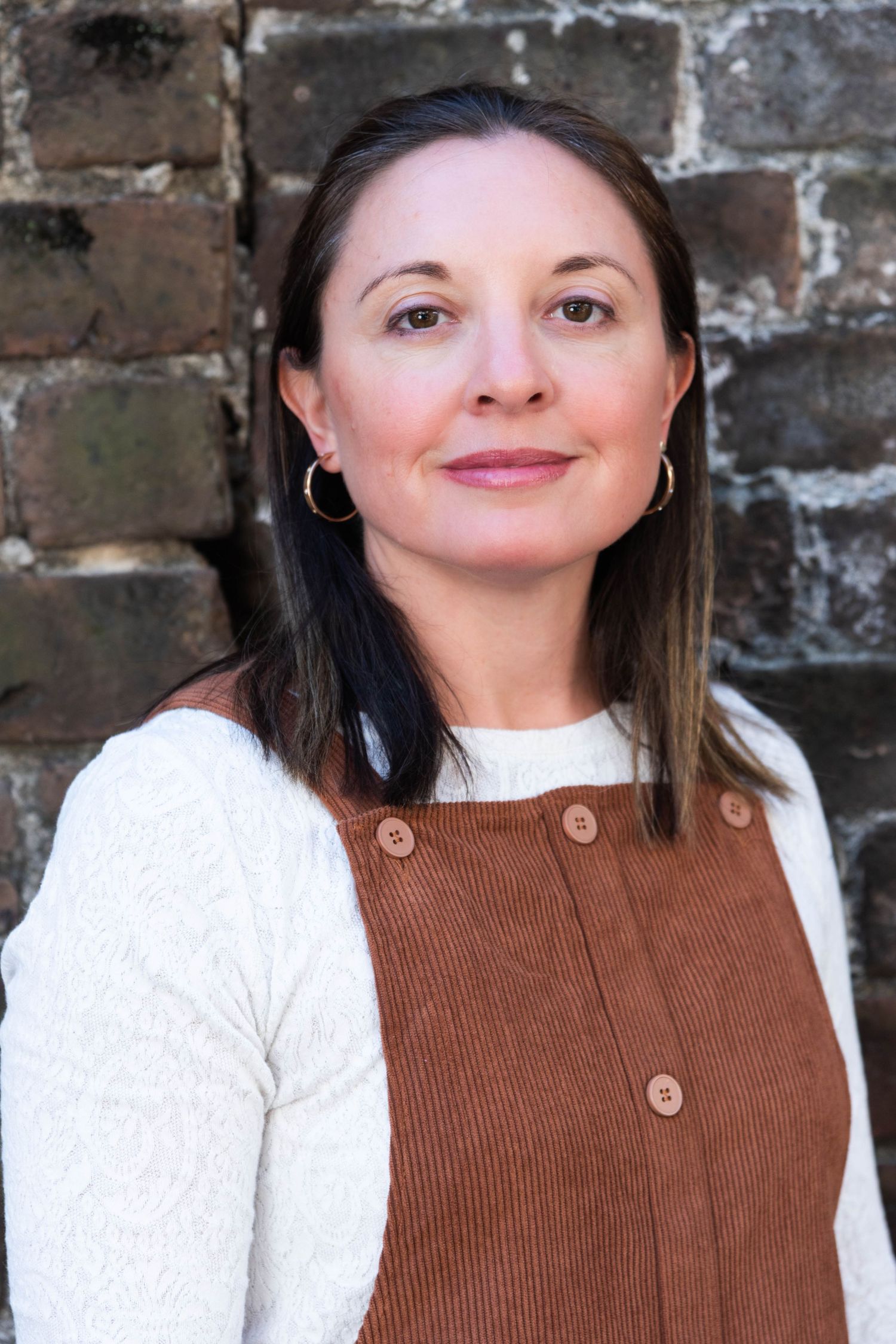 Person wearing a brown corduroy vest and white sweater poses against a rustic brick wall background.