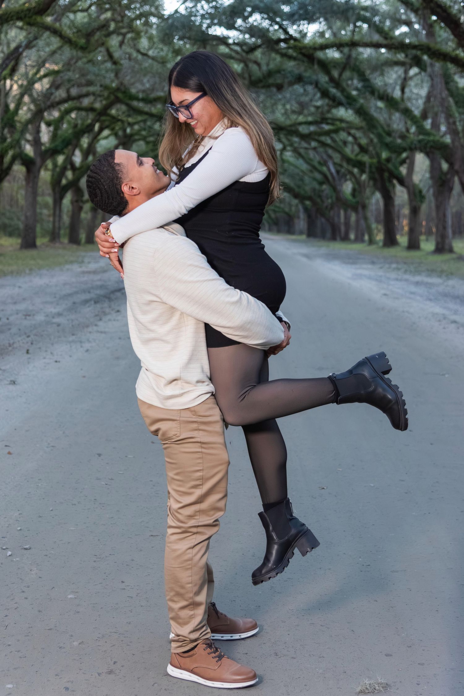 A man lifts a woman in a romantic embrace on a tree-lined path, she wears a black dress and brown boots.