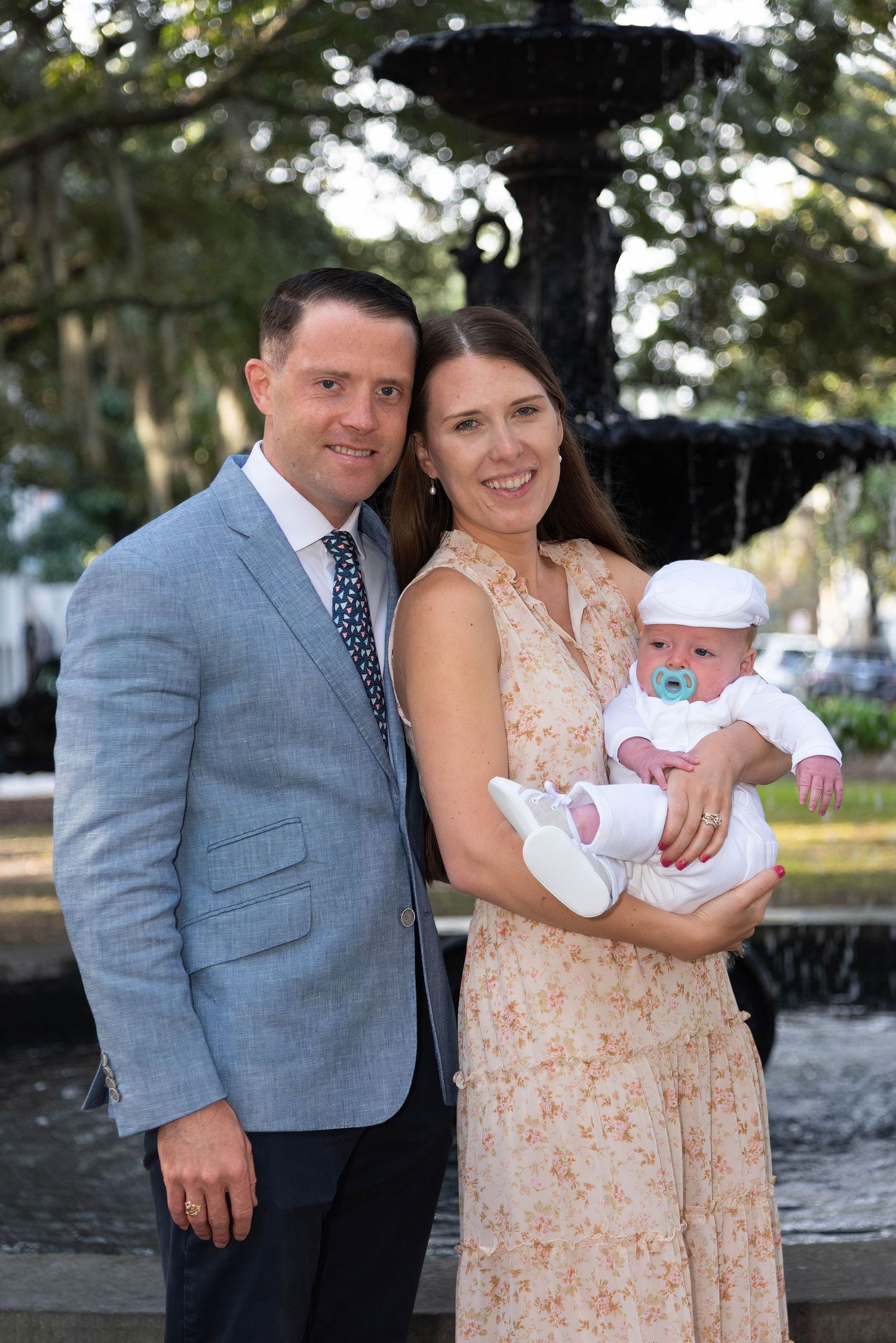 Family portrait in front of a garden fountain, wearing formal attire with a baby in white.