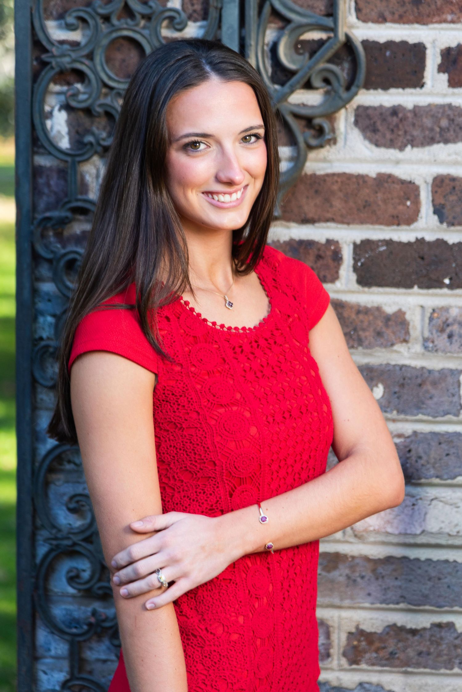 A woman in a bright red lace dress poses against a rustic brick wall with decorative iron scrollwork.