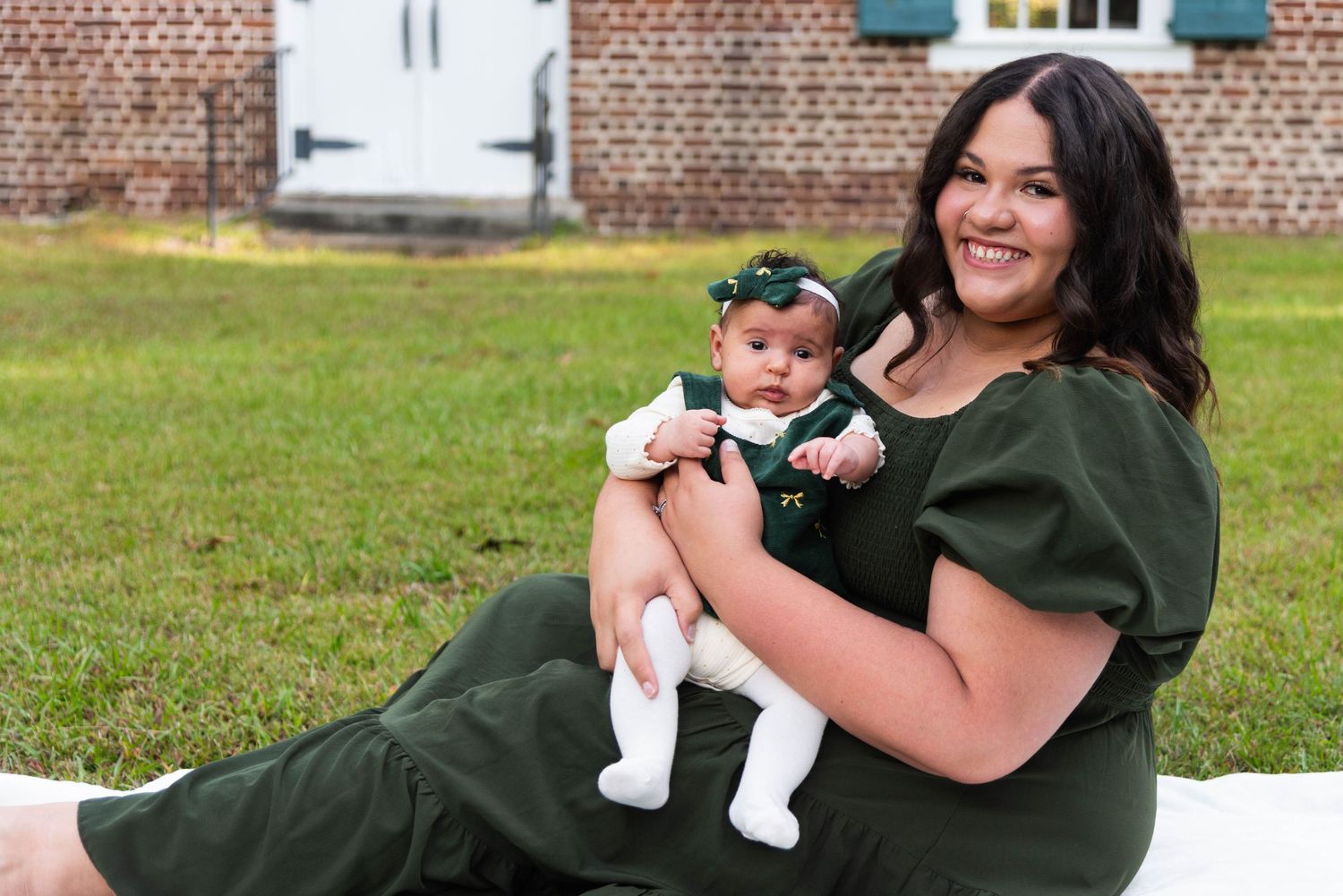 A woman in a green dress sits on grass holding a baby wearing a white outfit and bow near a brick building.