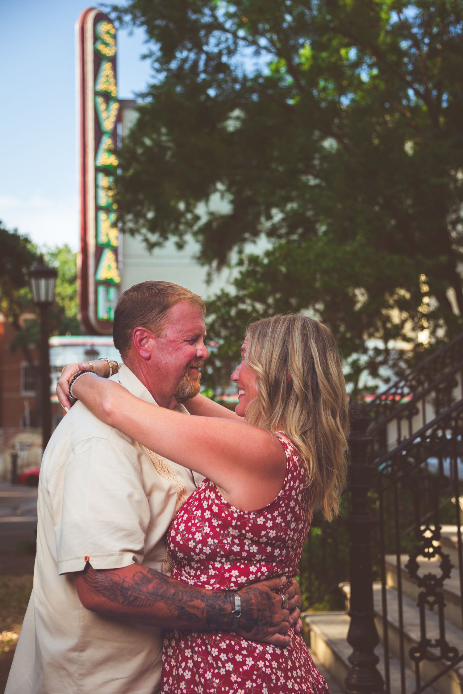 Couple embracing and smiling in front of a vintage Saveka neon sign outdoors.