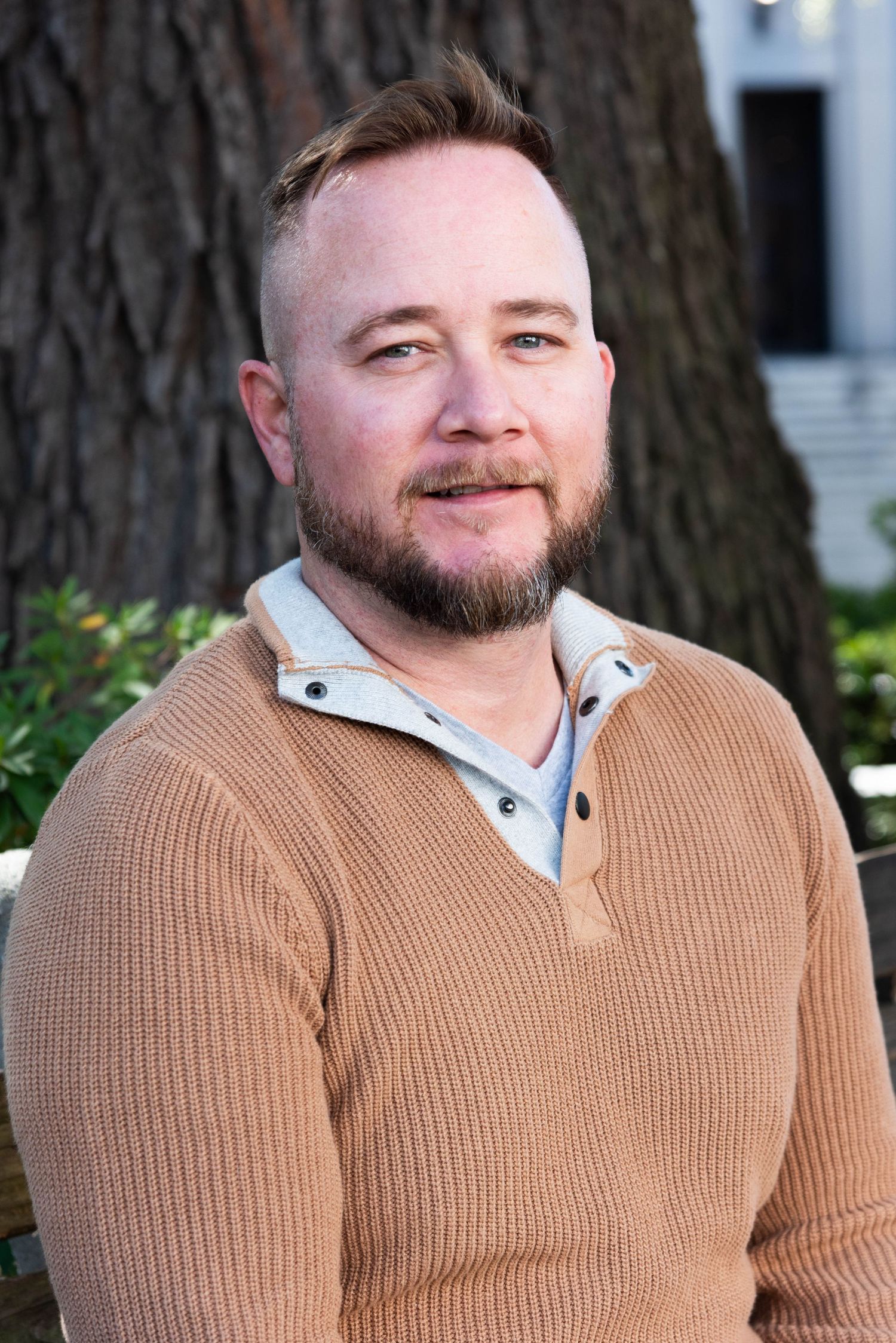 Professional headshot of someone wearing a brown knit sweater over a light colored button-up shirt outdoors.