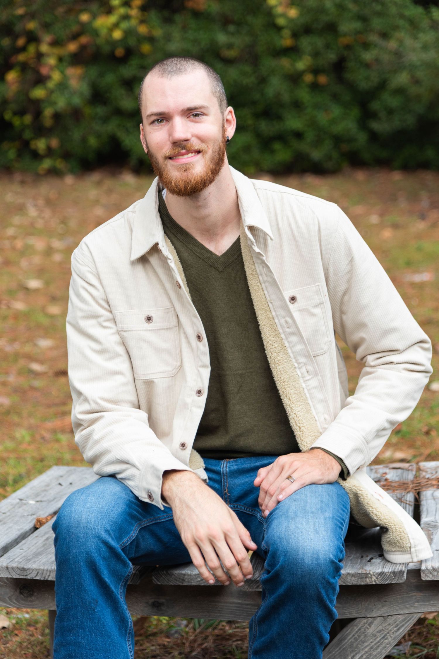 A person in a beige jacket and dark v-neck shirt sits casually on a wooden bench outdoors during autumn.
