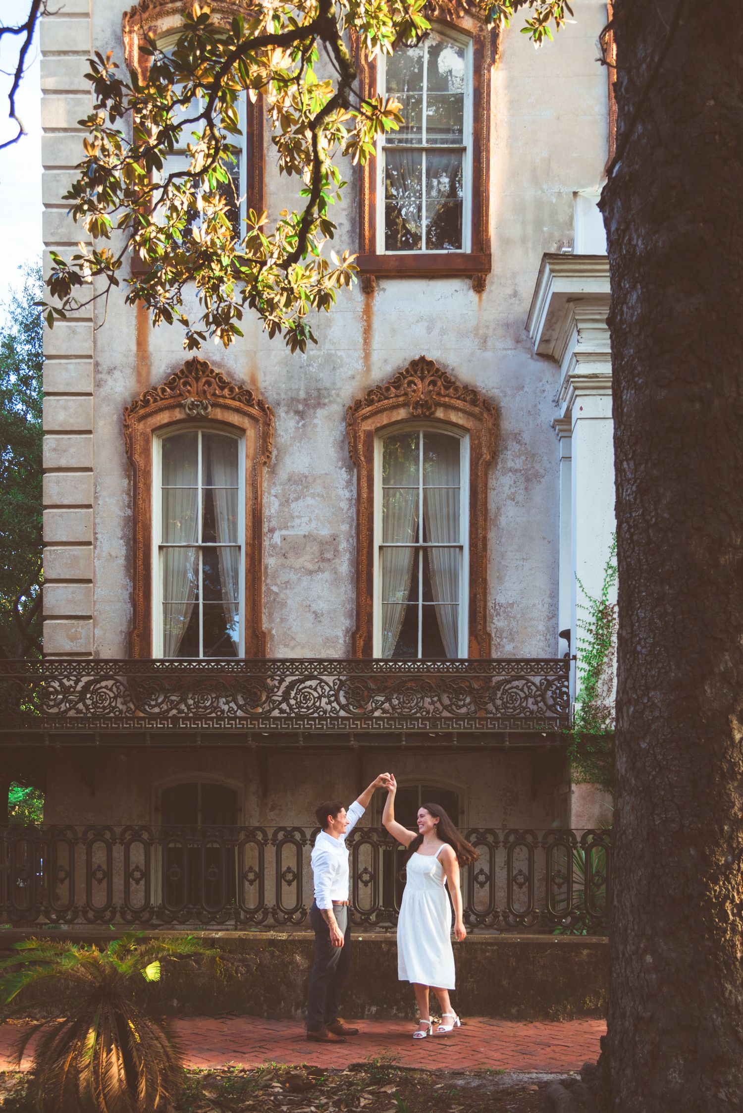 Couple dancing in front of a historic mansion with ornate windows and iron balcony railing surrounded by lush trees.