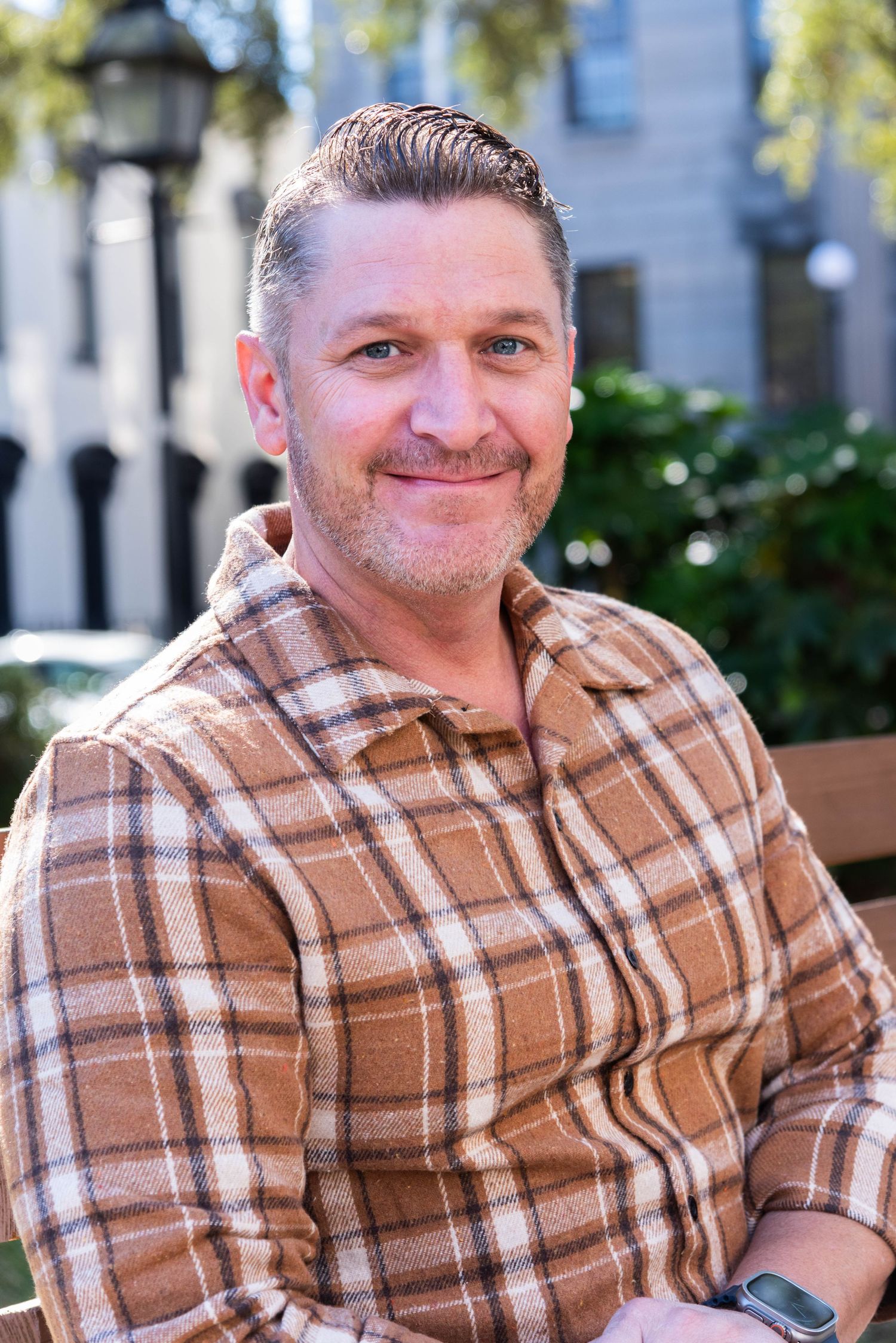 A person wearing a brown and white plaid flannel shirt sits outdoors on a sunny day with trees and buildings in the background.