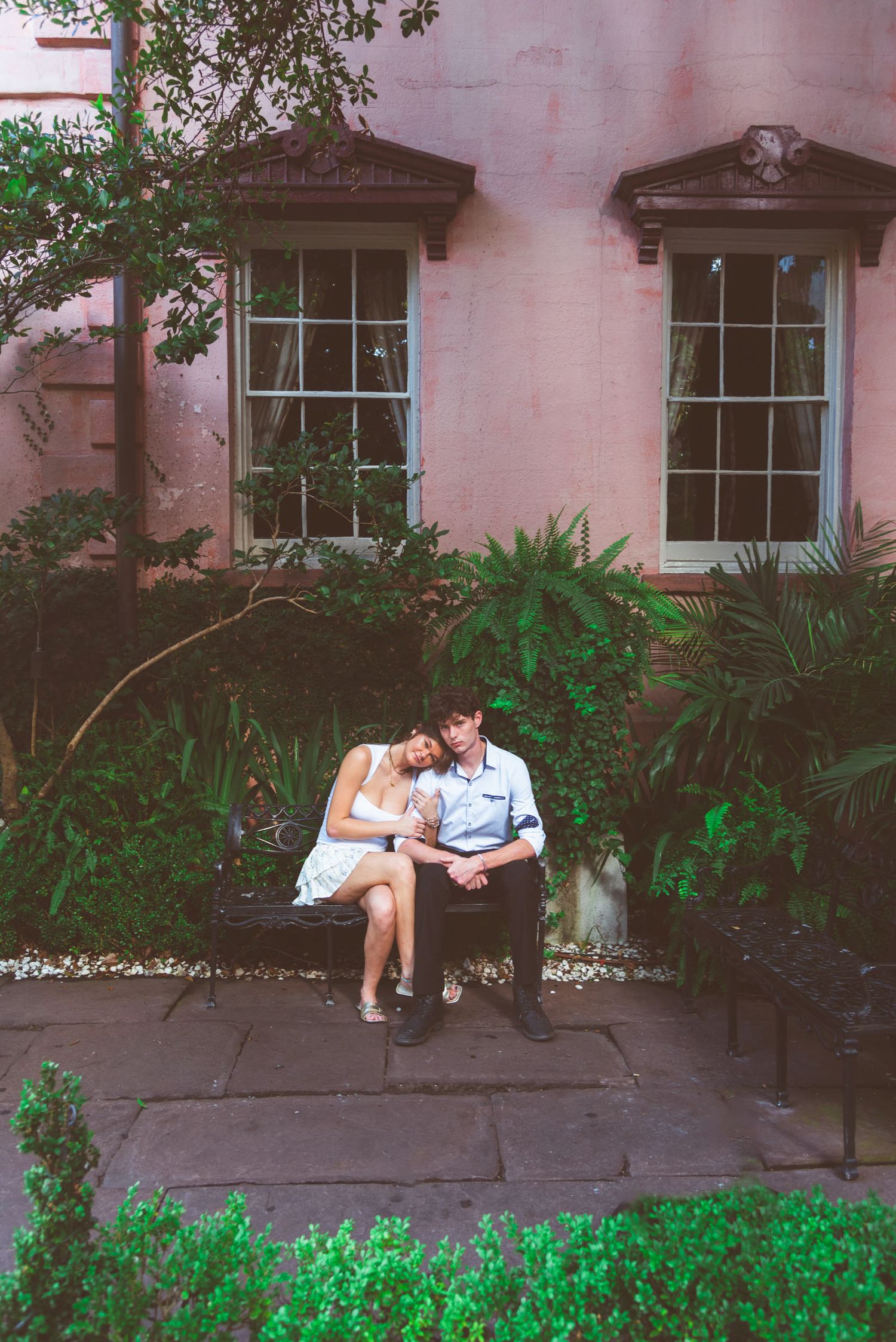 Couple sitting close together on a bench surrounded by lush greenery outside a pink building.
