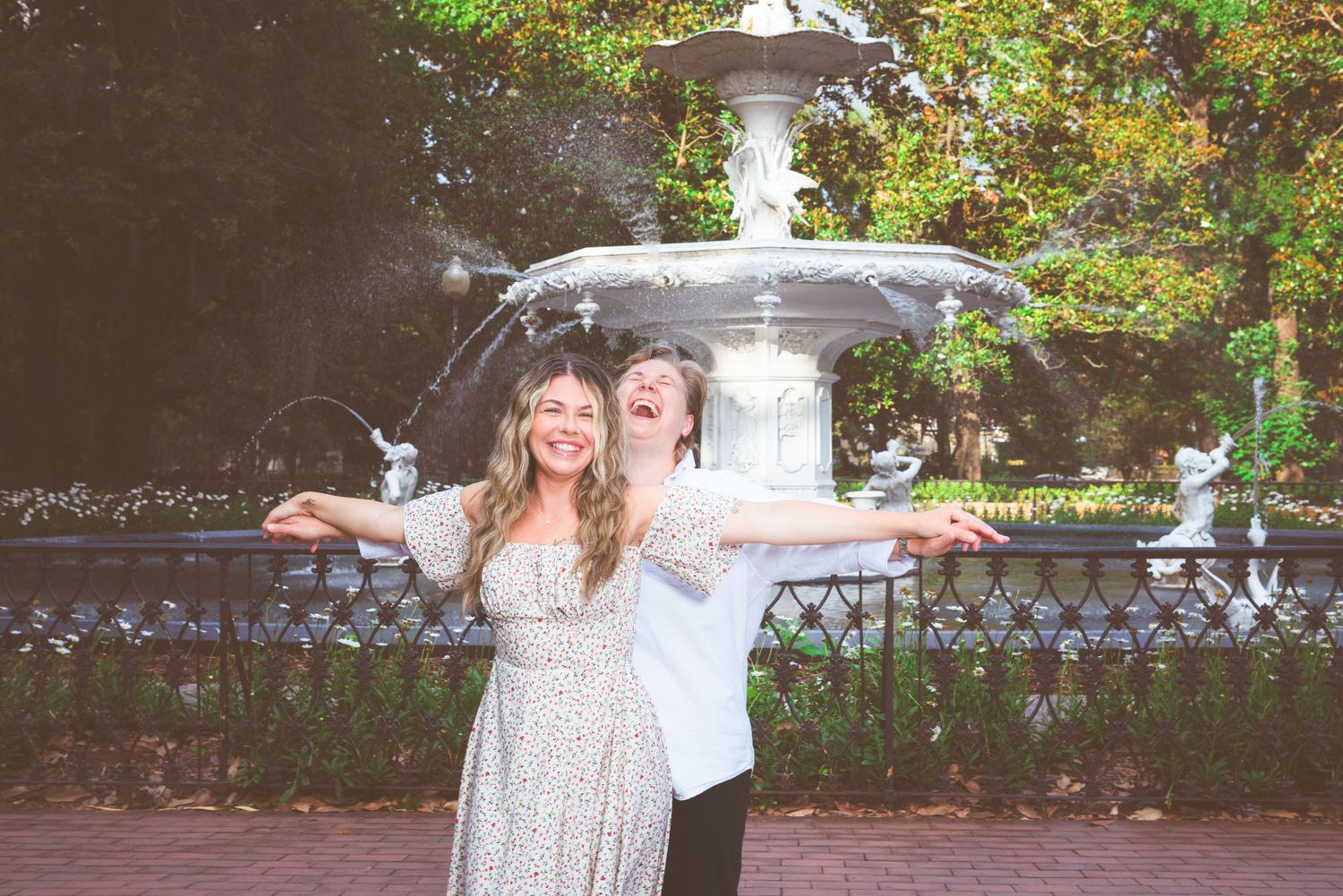 Two joyful women laughing with arms outstretched near a ornate fountain in a lush park setting.