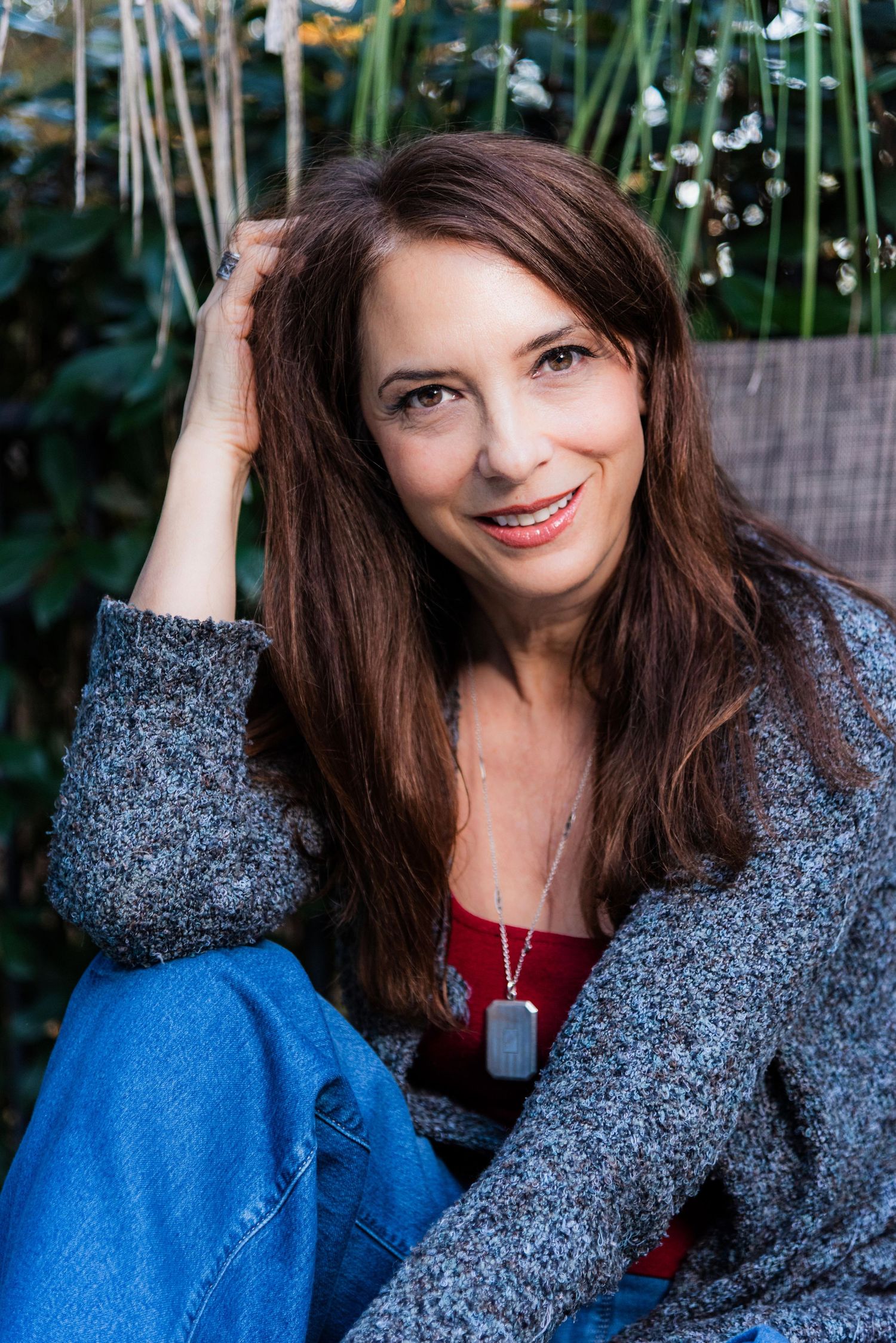 A person wearing a blue denim shirt and sparkly gray cardigan poses outdoors against a natural green background.