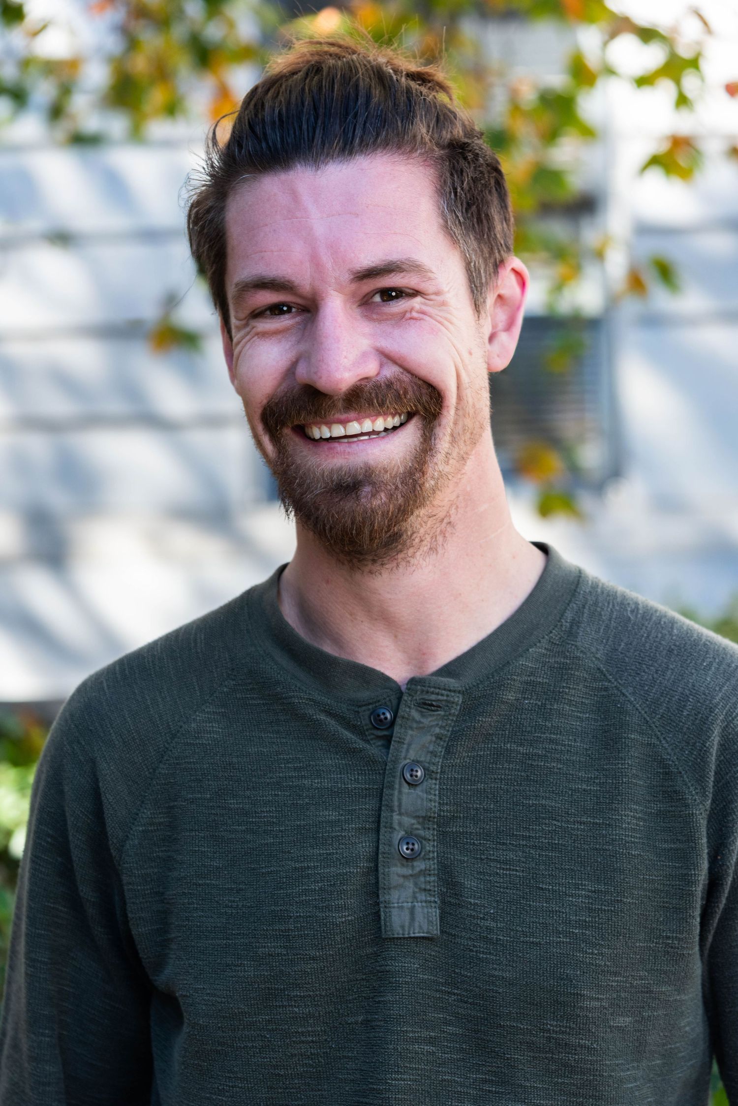 A person wearing a dark green henley shirt smiles warmly outdoors with autumn foliage in the background.