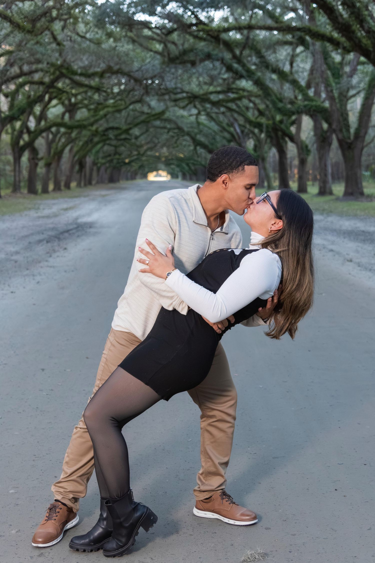 A couple in casual attire dances romantically on a tree-lined path, performing a graceful dip sequence.