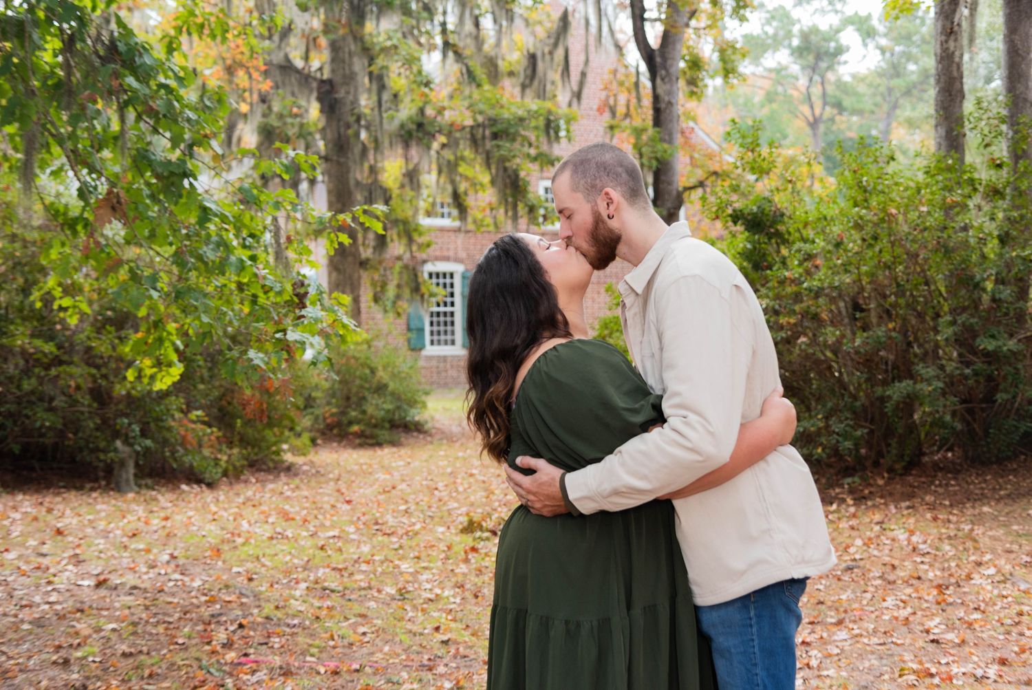 A romantic couple embraces on autumn grounds with fallen leaves and trees surrounding them in a natural setting.