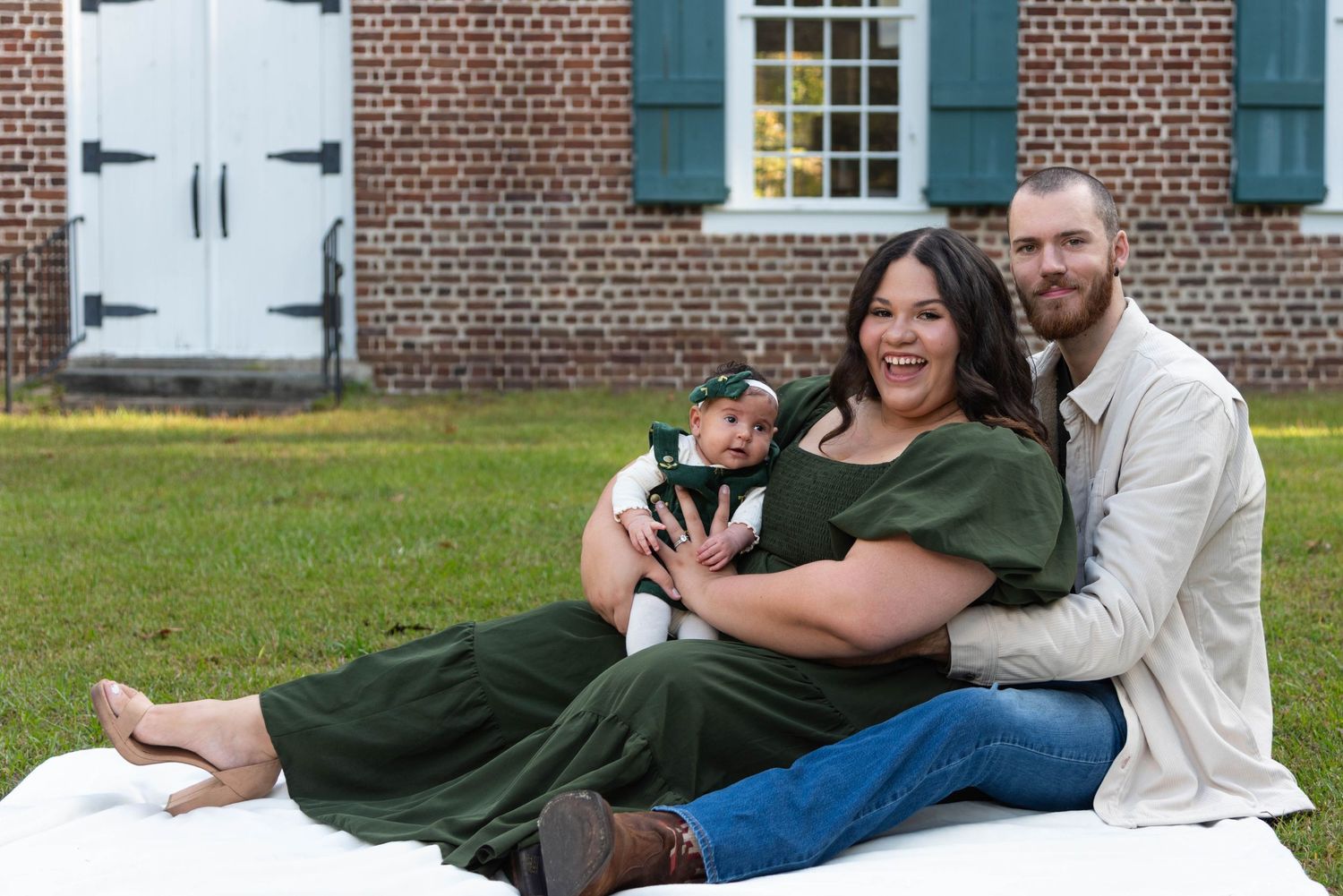 A couple relaxes on a blanket in front of a brick building with green shutters during an outdoor photo session.